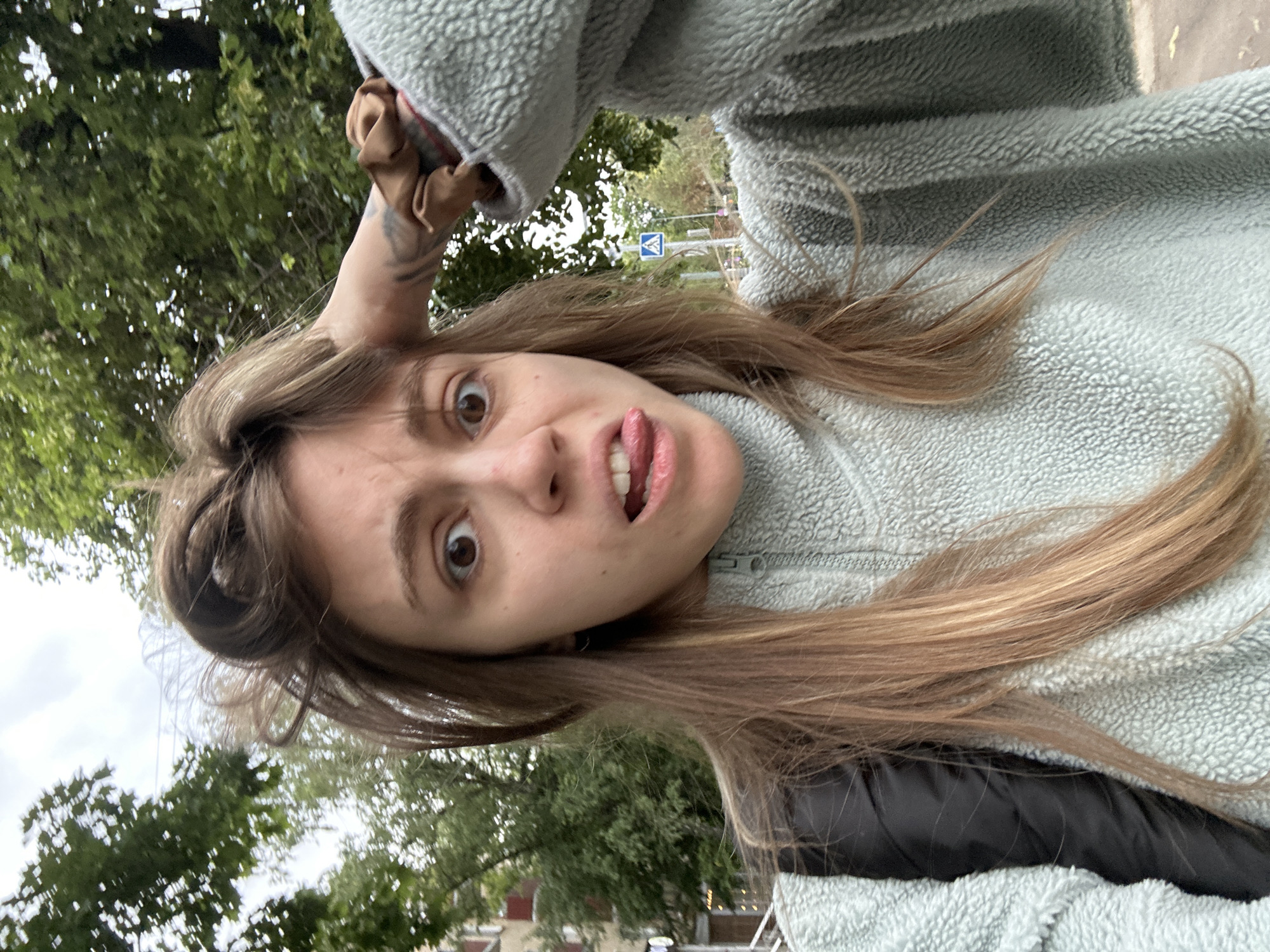A young woman with long, light brown hair sticks out her tongue and makes a surprised face at the camera. She is wearing a light blue fleece jacket with a zipper. Her hair is partially covering her face, and her left hand is visible touching her head. Behind her, green trees and a cloudy sky are visible. A blue and white road sign is also visible in the background. The image appears to have been taken outdoors during the day.