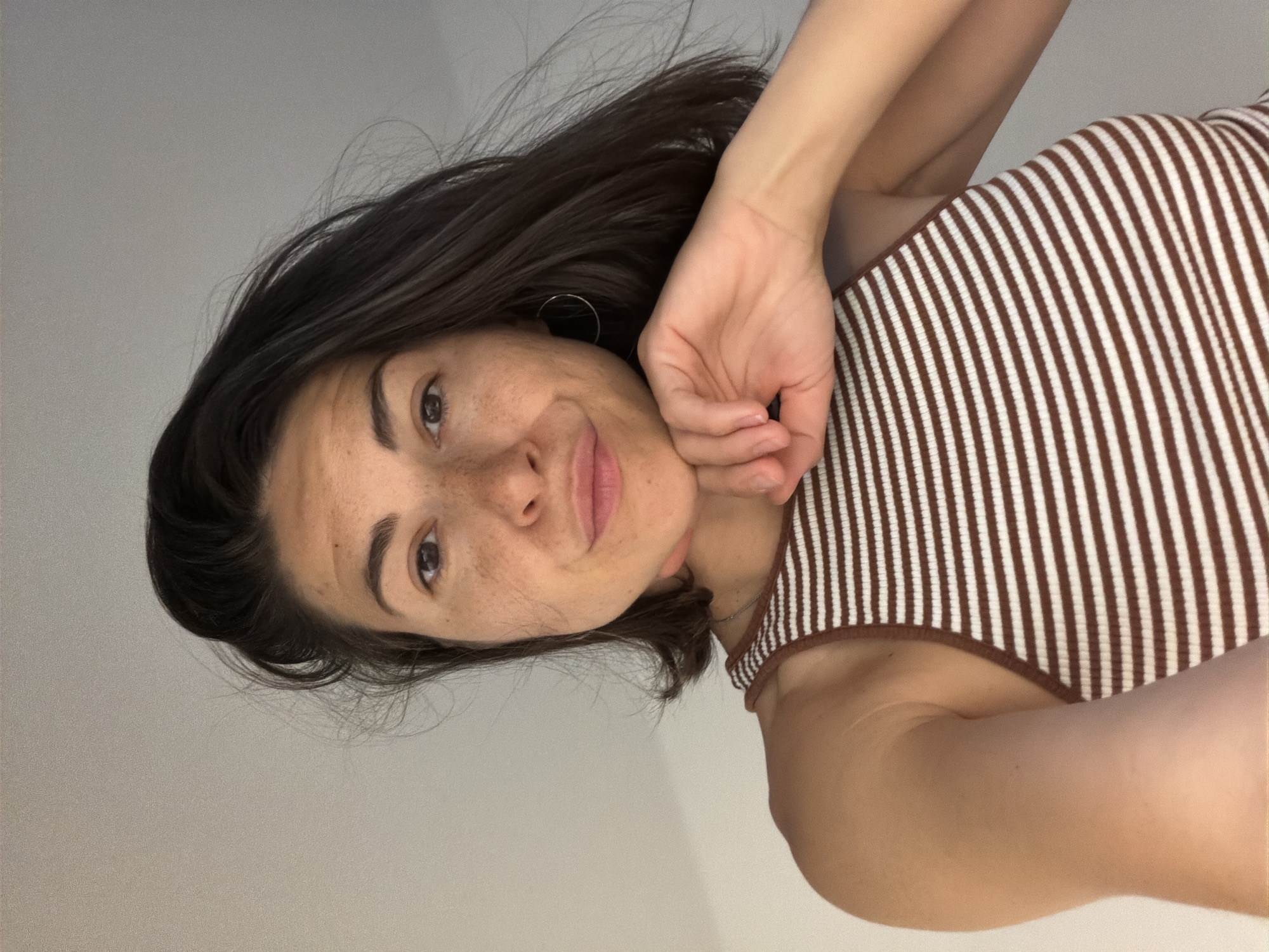 A woman with short, dark hair, freckles, and wearing a brown and white striped tank top is looking at the camera with a slight smile. Her left hand is resting on her chin. The background is a plain white wall. The image is oriented vertically. There is no information to indicate the location of the image, and therefore it is not possible to assert it was taken in Budva, Montenegro.