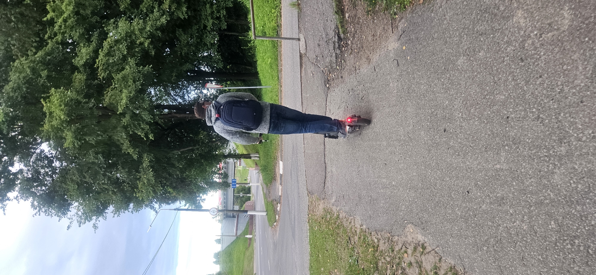A man wearing a gray hooded sweatshirt, blue jeans, and a dark blue backpack is riding an electric scooter on a paved path. The scooter has a red taillight illuminated. The path runs alongside a grassy area and appears to be a pedestrian walkway. In the background, there is a road with a bridge overhead. A traffic sign with the number 41 is visible on a pole near the road. The environment is green with trees and vegetation. The sky is cloudy. The scene is set in the city of Vitsebsk, Belarus.