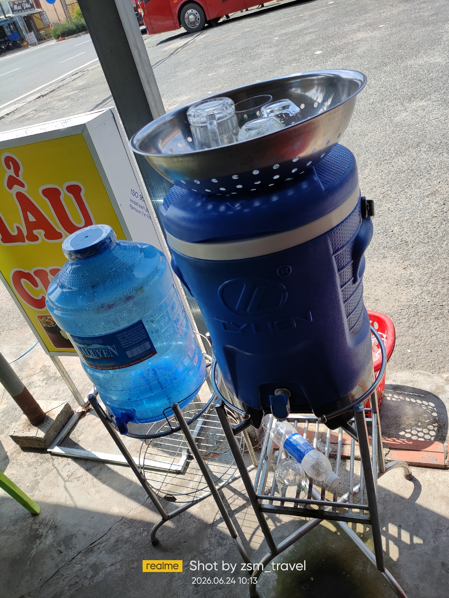 In Gò Dầu, Vietnam, an outdoor scene depicts a beverage stand situated on a concrete surface. The focus is on two large blue water dispensers and a metal stand supporting them. The dispenser on the left is a large, clear plastic bottle filled with blue-tinted water, condensation visible on its surface. The dispenser on the right is a larger, opaque blue container with a dispenser spout at its base. A stainless steel bowl containing several clear glasses rests on top of this dispenser. Below the dispensers, a metal shelf holds a partially filled plastic water bottle and another glass. A yellow sign with red lettering is partially visible behind the left dispenser, and a red bus is seen in the background on a paved road. The lighting suggests a sunny day, casting shadows on the ground.