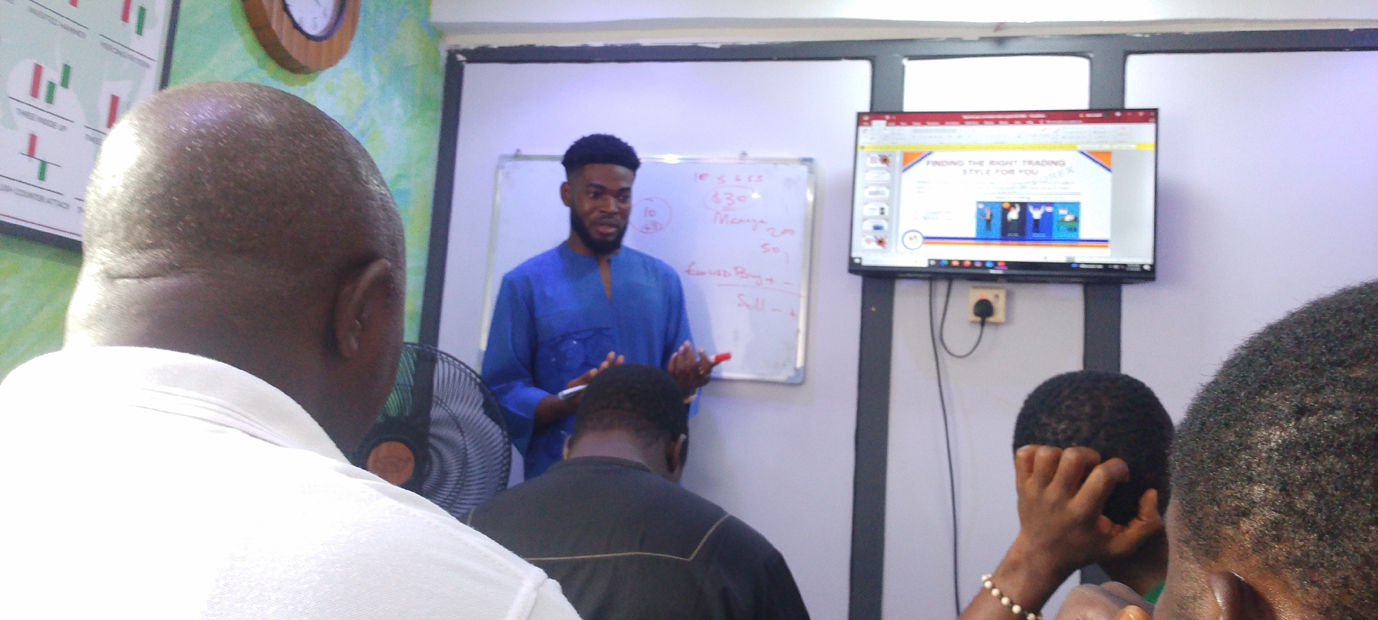 The image shows an indoor scene, potentially a training or workshop session, likely held in a room in Lokoja, Nigeria. A dark-skinned man in a blue tunic is positioned near a whiteboard, seemingly instructing or presenting to a group of attendees. The whiteboard contains scribbled notes related to calculations or financial terms. A monitor displays content titled "FINDING THE RIGHT TRADING STYLE FOR YOU" with graphic elements including portraits of men. Several individuals are seated, facing the speaker, with the backs of their heads visible. A fan is partially visible near the foreground. The room has white walls, and a clock hangs on a wall near a financial chart.