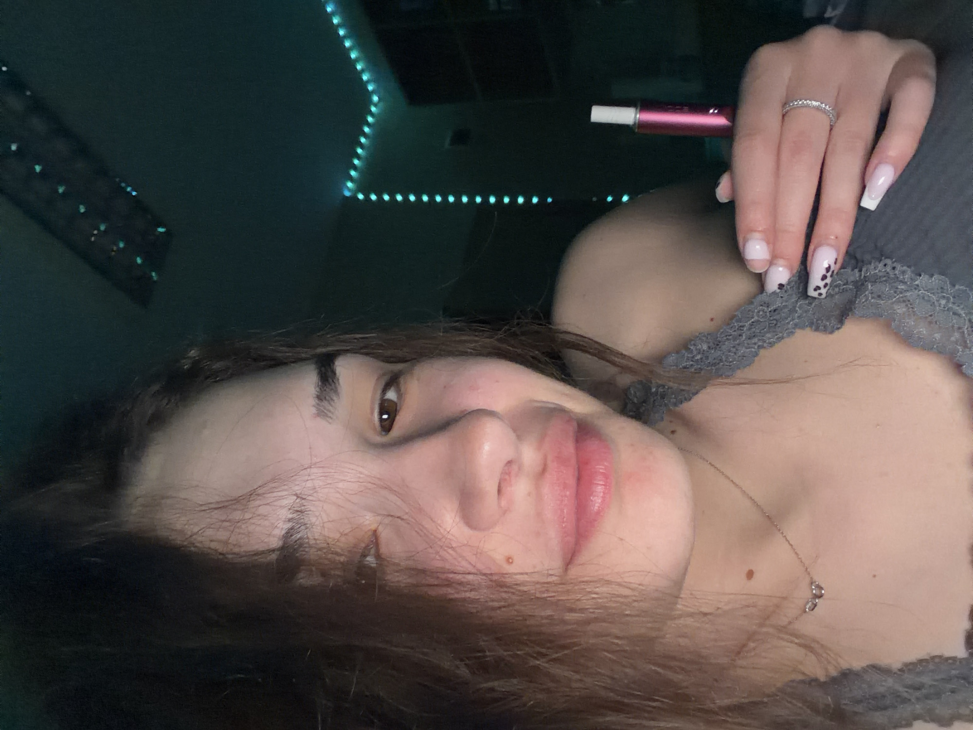 A young woman is lying down, facing the camera. She has brown hair, brown eyes, and fair skin. She is wearing a gray top with lace detailing. On her left hand, she has a silver ring on her ring finger and decorative false nails. She is holding a pink electronic cigarette. Behind her, there are string lights on the wall. The location cannot be determined from the image.