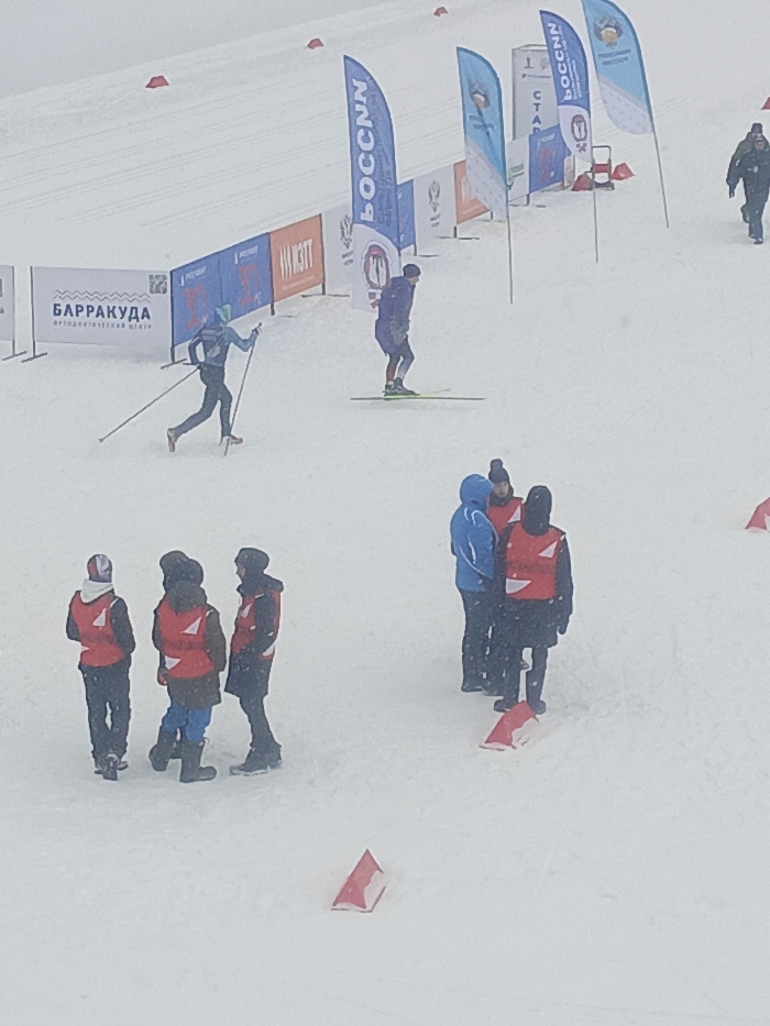 The image depicts an outdoor cross-country ski event or training session taking place on a snow-covered slope in Izhevsk, Russia. The environment is characterized by heavy snowfall or overcast conditions, resulting in reduced visibility.
In the upper-left, a cross-country skier in a teal-blue full-body suit and dark cap is actively propelling forward with ski poles. To their right, another skier in a dark blue/purple full-body suit stands stationary on skis, facing right. Further right, a person in dark winter attire walks away from the scene.
The background features several vertical banner flags, predominantly blue, some bearing the Cyrillic text "РОССИИ" or "РОССИЯ" with associated logos. Horizontal sponsor banners are visible, including a white banner displaying "БАРРАКУДА" and "ортодонтическая студия" (orthodontic studio) in Cyrillic. Red and white triangular marker cones are interspersed across the snow-covered area, defining boundaries or a course.
In the foreground, a group of seven individuals is clustered on the snow. Five of these individuals wear bright red reflective vests over dark winter jackets, indicative of event staff or volunteers. One of these wears a red helmet. The remaining two individuals are dressed in dark or blue winter jackets. They are observed standing, possibly conversing or monitoring the event.
