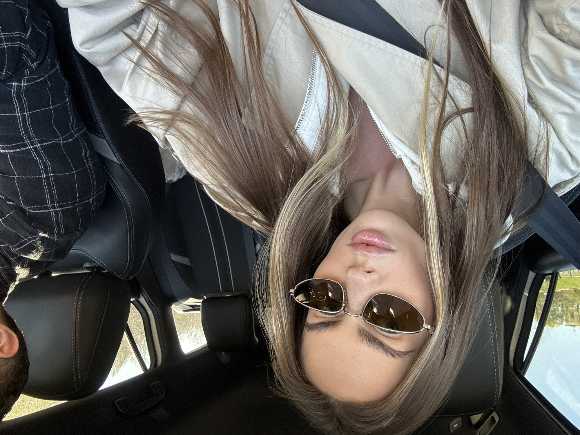 A person is in the passenger seat of a vehicle, wearing sunglasses and a beige jacket. The vehicle's interior is black leather. The person has light brown hair with blonde highlights. A seatbelt is visible. Another person wearing a black and white plaid shirt is partially visible in the driver's seat. Through the car windows, it appears to be daytime with greenery visible outside. There is no indication of this image being in Warsaw, Poland.