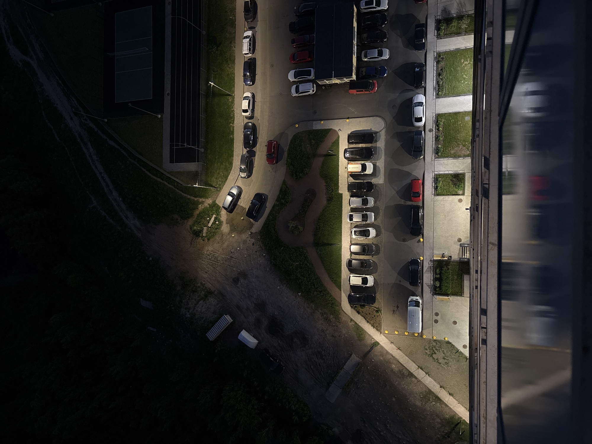 The image shows an aerial view of a parking lot and surrounding area in what is potentially Saint Petersburg, Russia. At night, light illuminates various sections, highlighting parked cars of different colors and sizes. A building with reflective windows is visible on the right side of the image. To the left of the parking area, there is a track, and a dirt path. Further in the background, some other buildings can be seen.
