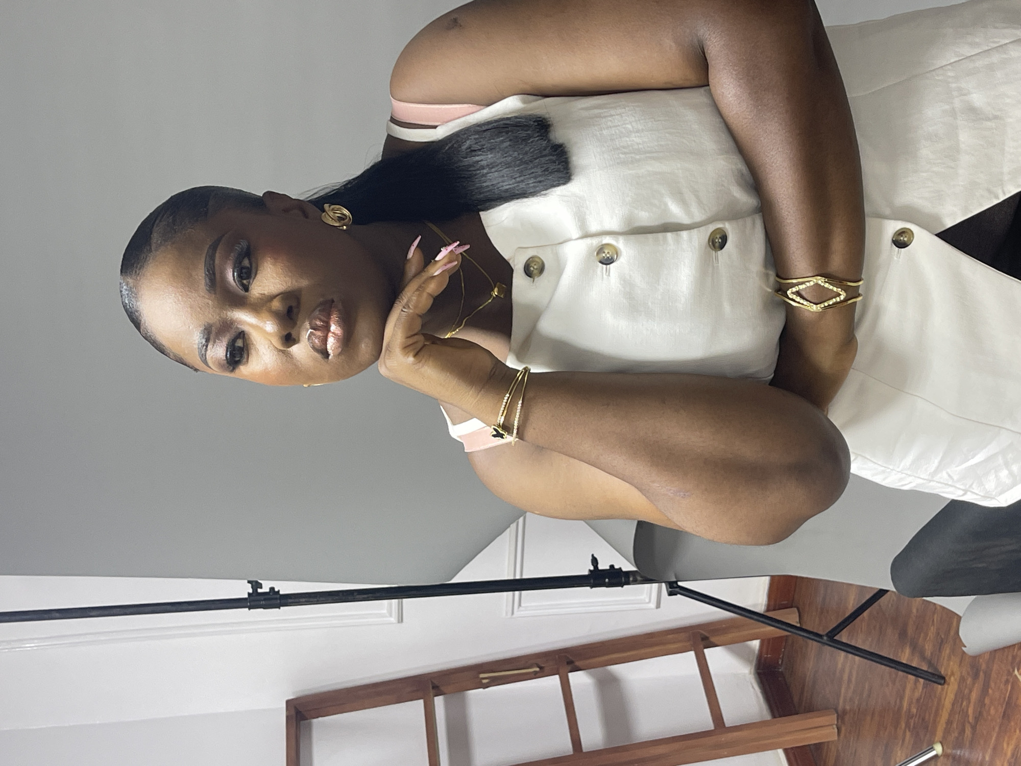 An indoor portrait captures a dark-skinned Black woman, likely in her late 20s or early 30s, posing directly for the camera. Her dark hair is slicked back, possibly in a bun or ponytail, with neatly gelled edges. She wears subtle eye makeup and glossy, metallic-toned lipstick, maintaining a composed and confident expression.
She is dressed in a light-colored, possibly off-white or cream, sleeveless button-down vest or top, featuring four visible light brown buttons on the front. A light pink strap, potentially from an undergarment, is visible on her left shoulder. Her attire is accessorized with striking gold jewelry: intricate knot-design earrings, a delicate necklace with a small square pendant, and multiple bracelets. Her right arm features a wide gold cuff bracelet embellished with a diamond-shaped pattern, along with several thin bangles. Her left hand, with long, light pink polished nails, is gently placed near her chin.
The setting appears to be a photography studio or a dedicated indoor space in Abuja, Nigeria. The background comprises a plain light grey wall, transitioning to a white wall section with decorative molding lower down. A black stand, likely for photographic equipment, is visible in the lower left of the frame. A dark wooden, multi-tiered shelf or ladder-like structure is also present in the lower right, resting on a polished wooden floor. The lighting is even, characteristic of an artificial setup, and there is no visible text in the image.