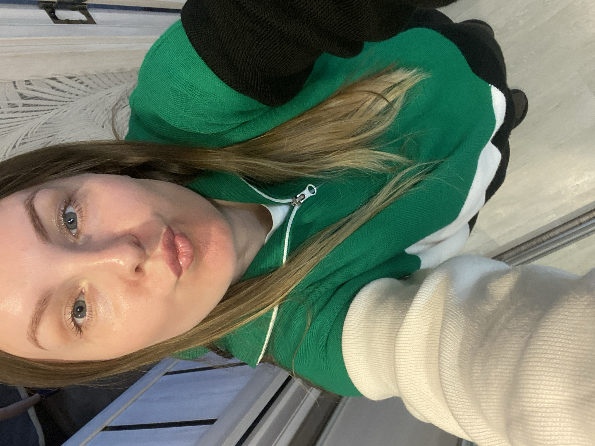 A fair-skinned female with light brown hair is wearing a multi-colored zip-up sweater. The sweater is green, black, and white. The location is Noviy, Russia. The woman is indoors.