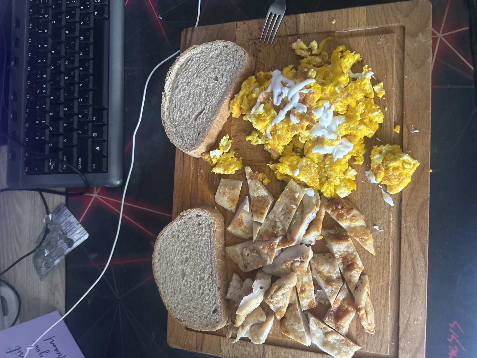 The image shows a wooden cutting board on a patterned surface. The board holds two slices of bread, a serving of scrambled eggs with white sauce drizzled on top, and sliced cooked chicken. A fork rests near the scrambled eggs. A computer keyboard and a medication blister pack are visible to the left of the cutting board. A handwritten note is partially visible in the lower-left corner. There is no information in the image that indicates location.