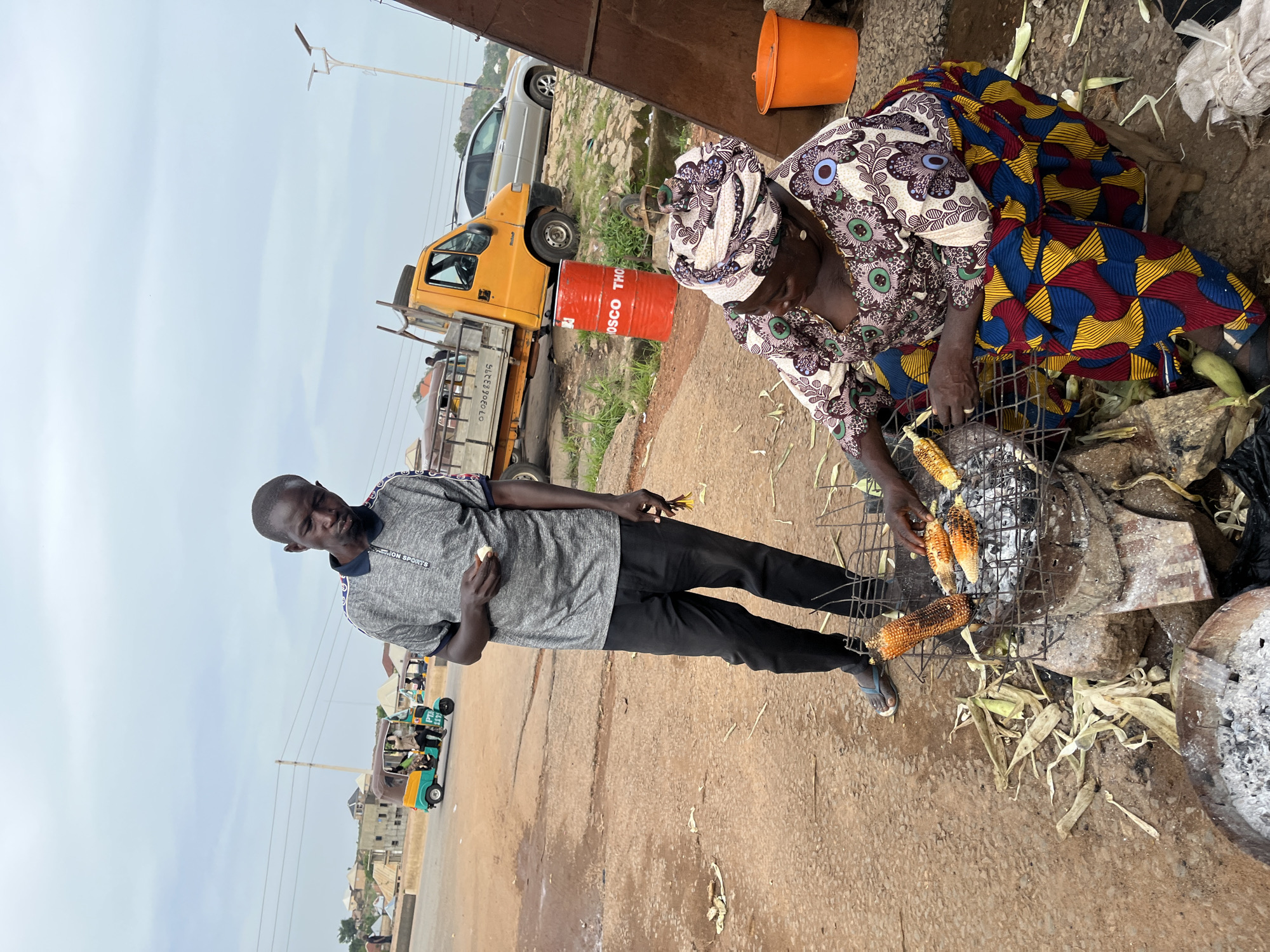 In Gura Top, Nigeria, a man and a woman are engaged in preparing corn. The man, dressed in a grey polo shirt and black trousers, stands with a piece of corn in his hand, looking down. The woman, wearing a vibrant, patterned dress and headscarf, kneels beside a charcoal grill, tending to several ears of corn being roasted. The grill is made of wire mesh and sits atop stones, with burnt corn husks scattered around it. In the background, a yellow truck, a silver car, and a colorful tricycle are visible, along with buildings and a cloudy sky, suggesting an outdoor market or roadside scene. An orange bucket and a red drum with writing are also present in the environment.