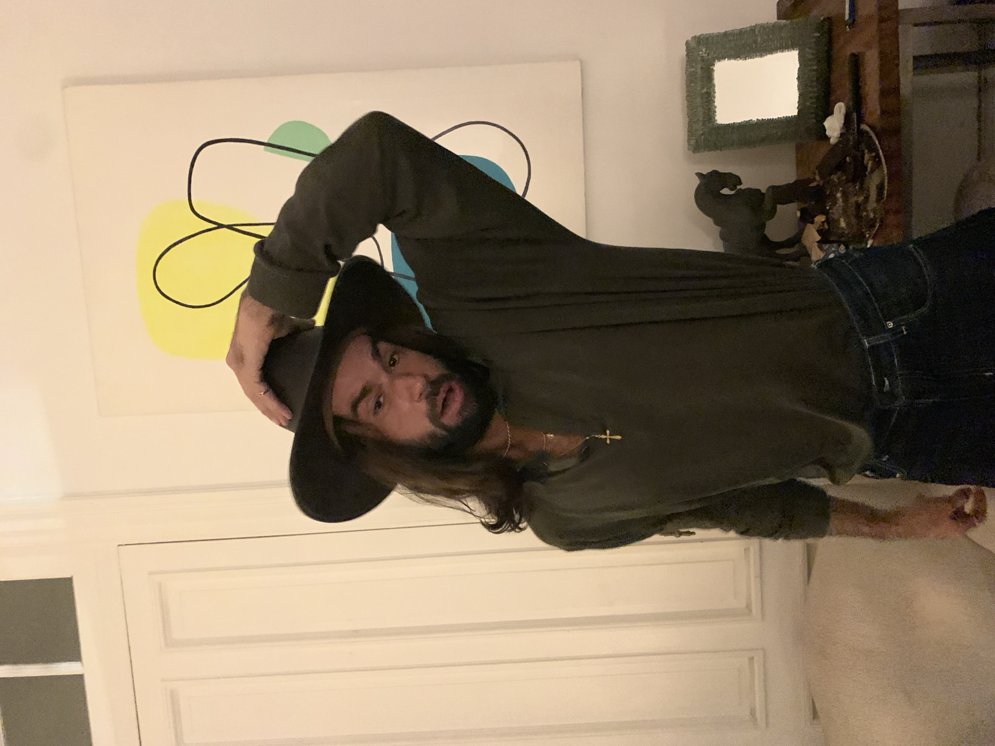The image depicts a man wearing a black hat, an olive green long-sleeved shirt, and dark wash jeans. He is holding the brim of his hat with his left hand. He has long brown hair and a beard, and he is wearing a gold necklace with a cross pendant. Behind him is a white wall with a painting that features yellow, green, and blue abstract shapes outlined in black. There is a white door in the background. To the right of the man, there is a dark-colored decorative object on a shelf. The image is taken indoors. The location of the photo is not known, so Buenos Aires, Argentina cannot be confirmed or denied.
