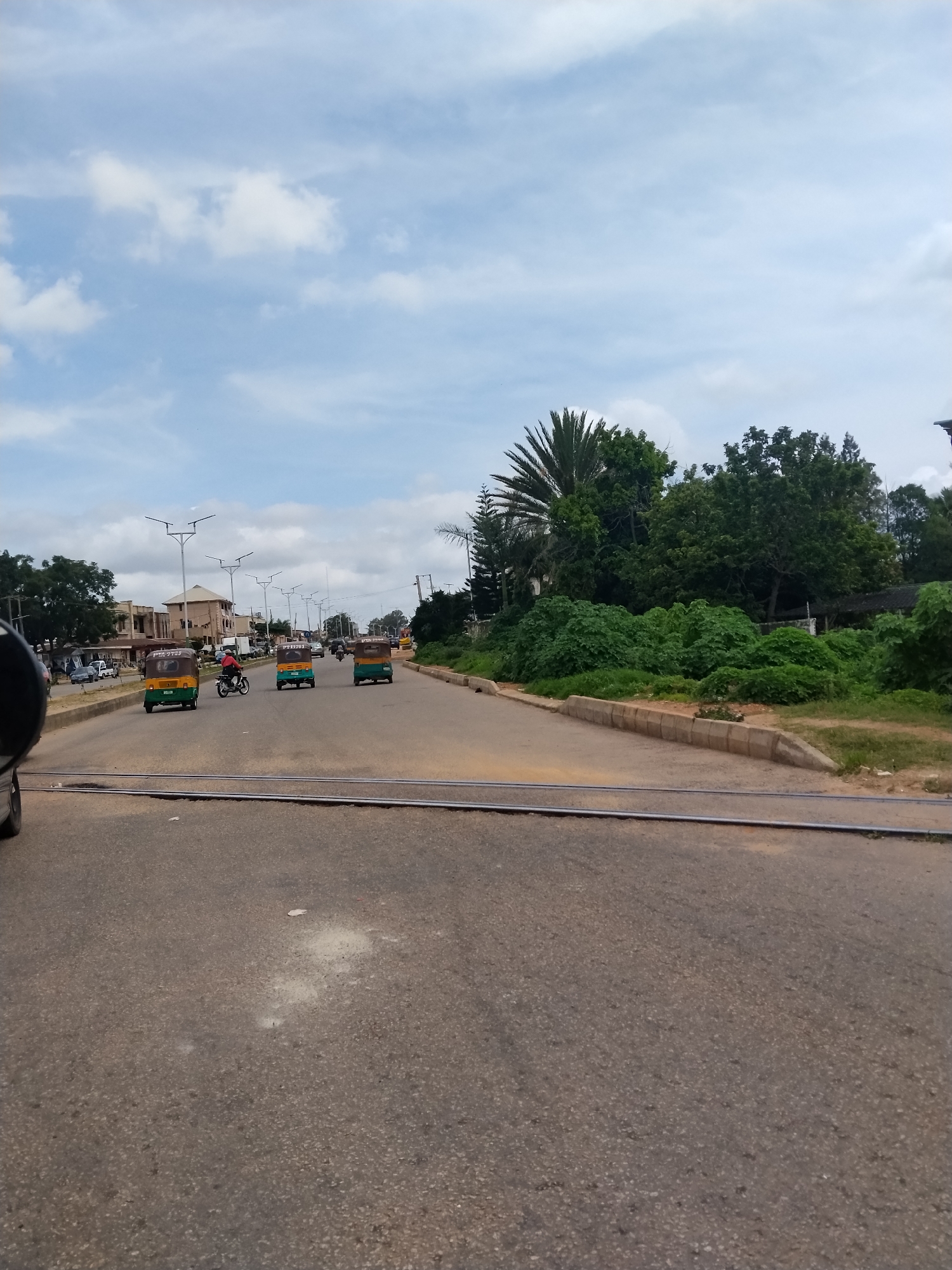 The image depicts a street scene in Jos, Nigeria, with a paved road running horizontally across the frame. A set of railway tracks runs across the foreground of the road. Several motorized tricycles, commonly known as Keke Napeps, are visible on the road, some moving and others stationary. A motorcycle with a rider is also present. Buildings and a variety of trees, including a palm tree, line the sides of the street. The sky is partly cloudy. The overall environment suggests an urban or semi-urban setting.