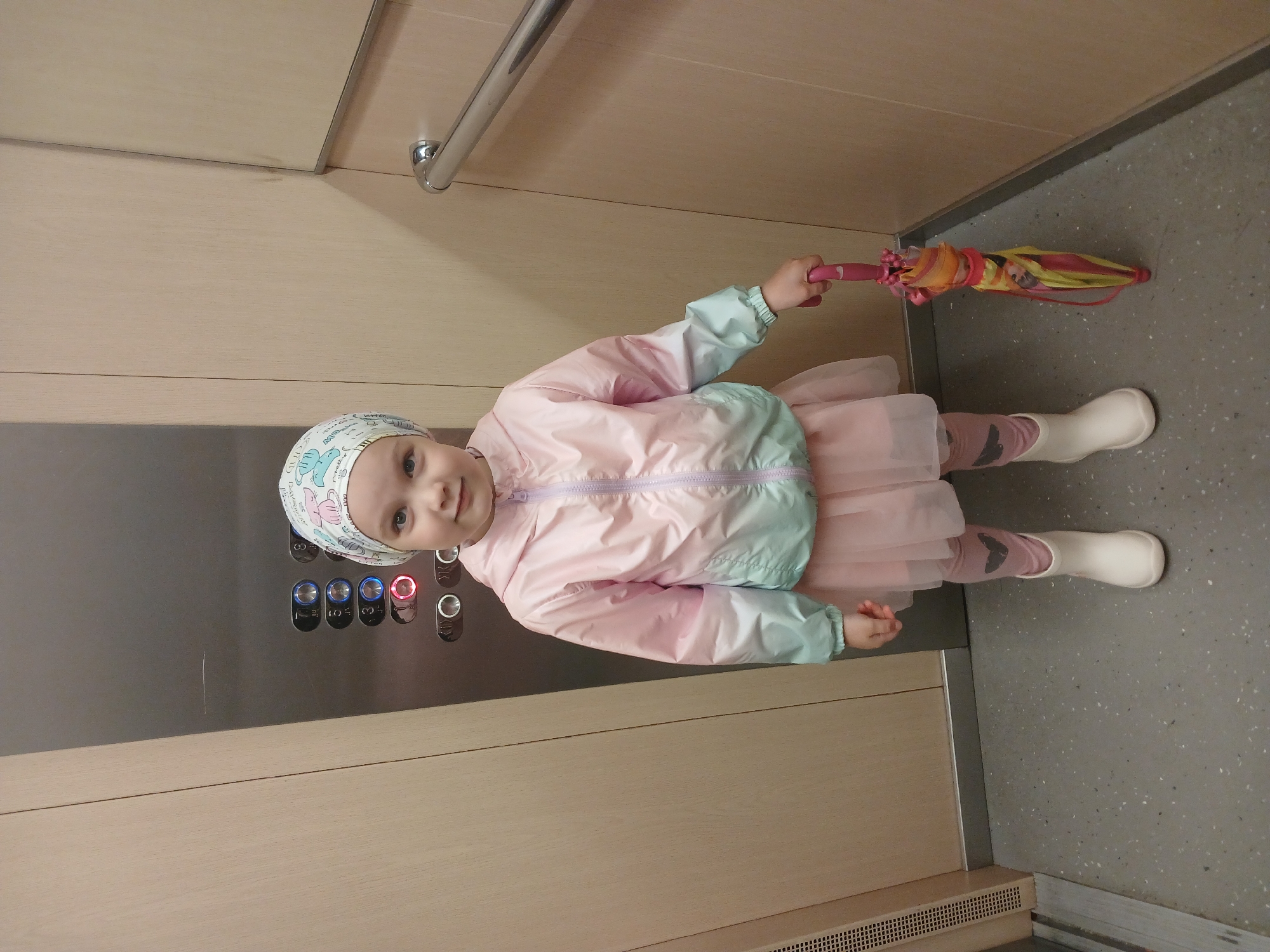 A young girl stands in an elevator, holding a closed, colorful umbrella. She is wearing a pink and mint green jacket, a pink skirt over pink leggings with a grey heart pattern, and white boots. A patterned headscarf covers her hair. The elevator walls are wood-paneled on the right and a dark metal panel with illuminated buttons on the left. The floor is speckled grey. The elevator buttons are illuminated red and blue, indicating floors 1 and 3 are active. The scene is likely within a residential or commercial building in Yekaterinburg, Russia, as suggested by the style of the elevator and building materials.