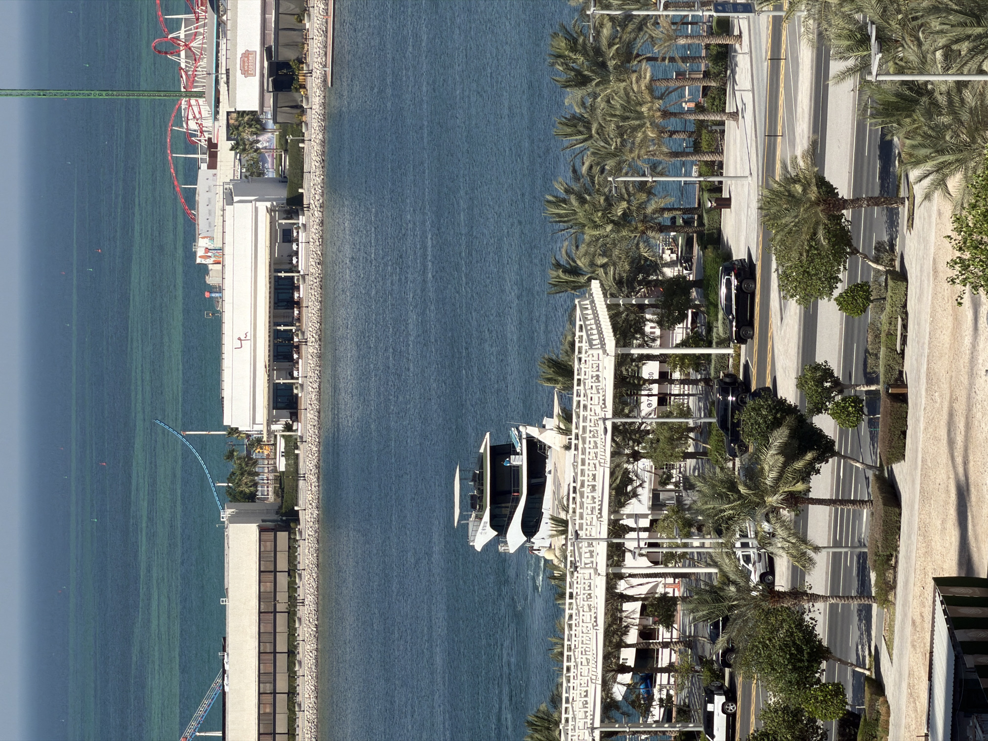 This is an aerial, rotated image of a waterfront promenade. The scene is divided into the sea on the left and a developed area on the right, which includes a road, buildings, and vegetation.
The sea is a vibrant turquoise, with distinct patterns indicating depths or underwater structures. A few buoys are visible on the water's surface. On the developed side, a coastal walkway or promenade is lined with palm trees and low-lying buildings. A roller coaster track, painted red and white, is a prominent feature in the upper left portion of the image. Further down the coast, a white structure resembling a modern building or a boardwalk with white railings is visible, and several white boats are docked. The road running parallel to the coast has multiple lanes and is marked with yellow lines. Several cars are parked or driving on the road, including a black SUV and a white van. The area is well-maintained with manicured trees and bushes.
The time of day appears to be daytime, with bright sunlight casting shadows. The weather seems to be clear and sunny. There is no explicit indication of the location, but the presence of palm trees and the style of architecture suggest a warm, possibly coastal or resort area. No people are clearly visible in the image.
Visible text includes a partial sign on a building that reads "30" and another that appears to be a license plate or building number starting with "07".