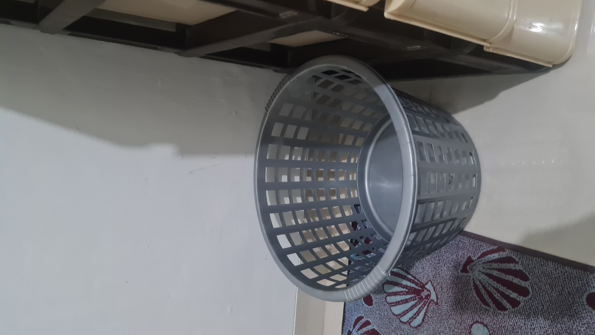 The image depicts a grey plastic laundry basket placed on a floor with a patterned rug. The rug has a repeating seashell motif in shades of red and blue on a grey background. To the right of the laundry basket, a dark brown shelf unit with beige drawers is visible. The wall behind the basket is plain and white, with some shadows cast by an unseen object. The scene appears to be indoors, likely in a domestic setting. The lighting is moderate, suggesting it could be daytime with artificial light. There is no indication of the specific location within Bandar Lampung, Indonesia, beyond the general context of it being an indoor environment.