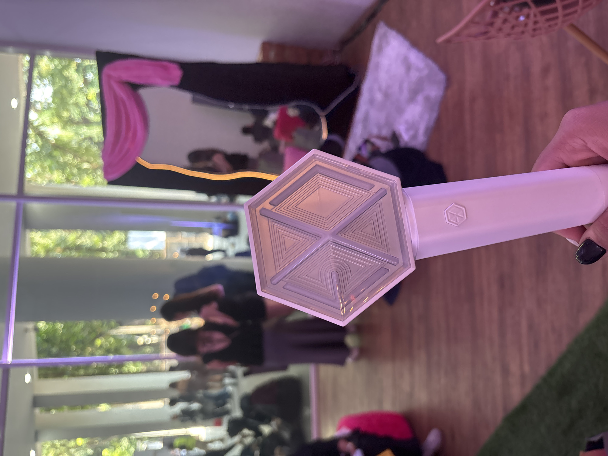 The scene depicts an indoor event space in Central Jakarta, Indonesia. In the immediate foreground, a person's left hand with dark-painted fingernails holds an illuminated white hexagonal K-pop lightstick, identified as an EXO-L official lightstick. The lightstick's head features an intricate geometric pattern of interlocking triangles and diamonds, emitting a subtle purple glow. The handle bears a small hexagonal EXO logo.
The floor transitions from a strip of artificial green turf on the lower left to a polished dark brown wooden surface that covers most of the visible ground.
In the midground, slightly out of focus, two women stand conversing. The woman on the left wears a dark sleeveless top and a long grey skirt, with her back partially turned towards the camera. The woman on the right has long dark hair and wears a dark top, facing her companion.
To the right, a large rectangular mirror is positioned, framed by black material and adorned with draped pink fabric across its top edge. A vertical yellow neon-like light strip illuminates the left side of the mirror's frame. A white, textured rug lies on the floor in front of the mirror. A bright pink beanbag chair is partially visible on the far left.
The background reveals an expansive, modern event space with large, floor-to-ceiling windows, separated by light-colored architectural frames. Beyond the windows, dense green foliage from trees is visible, indicating an outdoor garden or park setting bathed in natural daylight. Numerous blurred figures of other event attendees are visible throughout the background, both inside and through the windows. The interior is illuminated by a mix of ambient purple artificial light and natural light filtering through the windows, creating a lively social atmosphere.