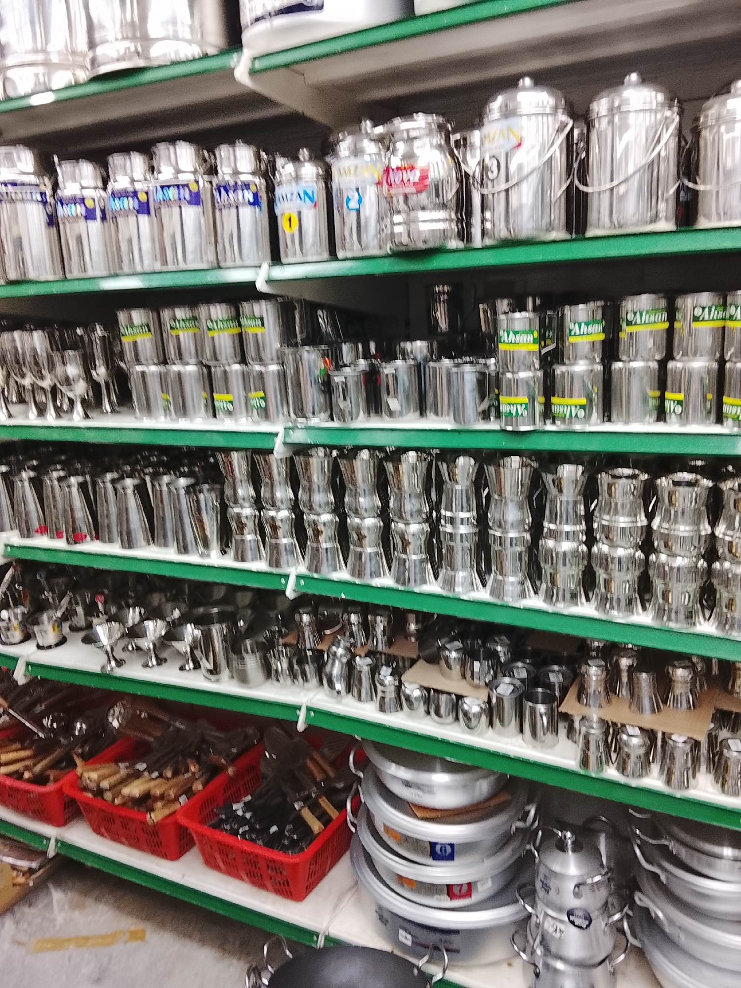 The image depicts a retail setting, specifically a shop in Gulgasht Colony, Pakistan, displaying kitchenware.
The scene features shelves stocked with various stainless steel and aluminum kitchen utensils and cookware. The top shelves primarily hold tall, cylindrical containers with lids, some with handles. Below, are shelves packed with smaller utensils, including metal cups, pots, and other cooking tools. The lowest shelves contain red plastic baskets filled with wooden-handled items and stacks of aluminum pots and pans. The lighting is artificial, illuminating the polished metal surfaces of the items.