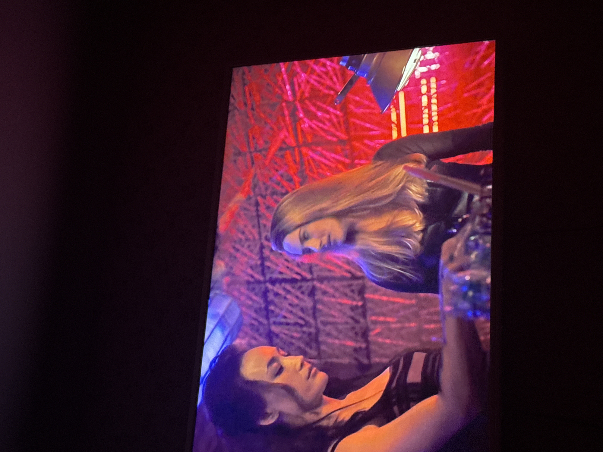 Two women are displayed on a screen. One woman has dark hair and is wearing a tank top. The other woman has blonde hair and is wearing a dark long-sleeve shirt. The background is red with a pattern. There is a lamp shining on the women. The location is indiscernible.