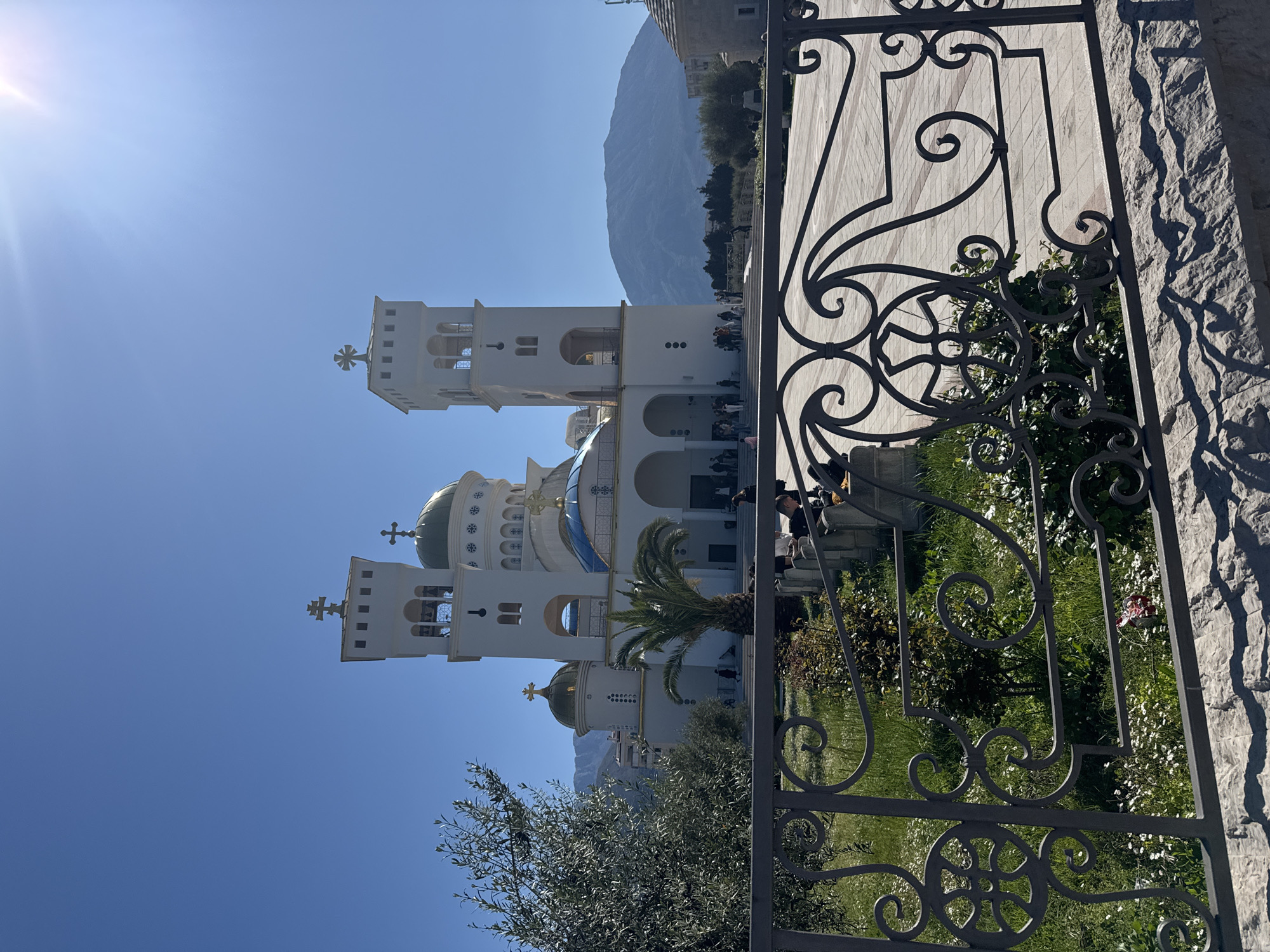 The image captures a view of the Church of St. Jovan Vladimir in Bar, Montenegro, seen through a wrought-iron fence. The church is a prominent white structure with multiple towers, topped with crosses and green domes. People are seen around the base of the church. In the background, a mountain range is visible under a clear blue sky. The foreground features the decorative iron fence, adding depth to the composition. A stone wall is seen to the right of the fence. A tree and some greenery are also visible. The scene appears to be taken during daylight.