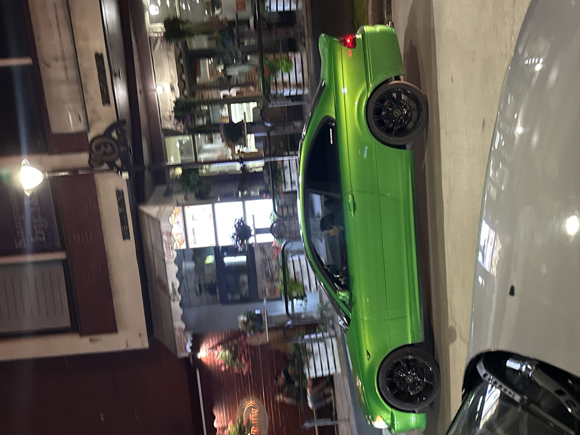 In Veliko Tarnovo, Bulgaria, a vibrant green BMW coupe is captured in profile at street level, parked on asphalt. The car features black rims and a distinct spoiler on its rear. In the background, a building facade with maroon and cream accents is visible, housing several businesses. One establishment has a red and white striped awning, and the text "Coca-Cola" is discernible. To the left, a street lamp with a glowing light fixture stands prominently. A glimpse of another vehicle's side, with a black trim and a reflective surface, is present in the lower portion of the frame. The scene appears to be set during the evening or night.