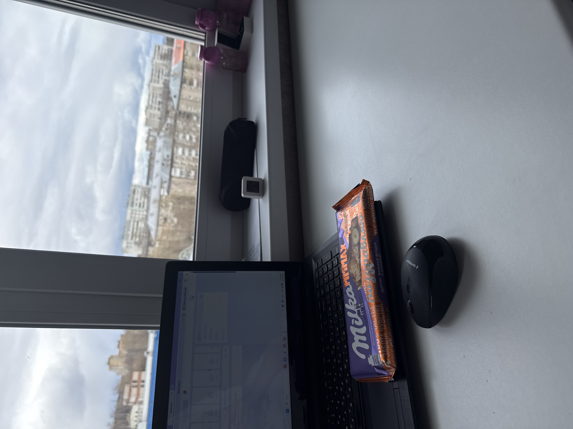 The image shows a desk next to a window with a view of Perm, Russia on a cloudy day. A black laptop computer is open on the desk. A Milka MMMax chocolate bar rests on the keyboard. A black Gembird wireless computer mouse sits to the right of the laptop. On the windowsill are several perfume bottles, a black pouch, and a digital thermometer displaying the time and temperature. The laptop screen displays a webpage related to VKontakte.