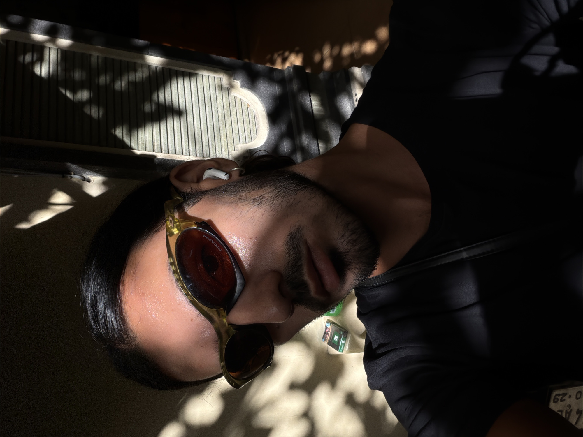 A man with dark hair and a beard is lying down, facing the camera. He is wearing gold-rimmed sunglasses with dark lenses and an earbud in his left ear. He is dressed in a black shirt. Patches of bright sunlight and shadow fall across his face and clothing. There are small, rectangular objects near his neck. The background includes a wall with vertical slats and dappled light and shadow. A license plate is partially visible. The location is North Kuta, Indonesia.