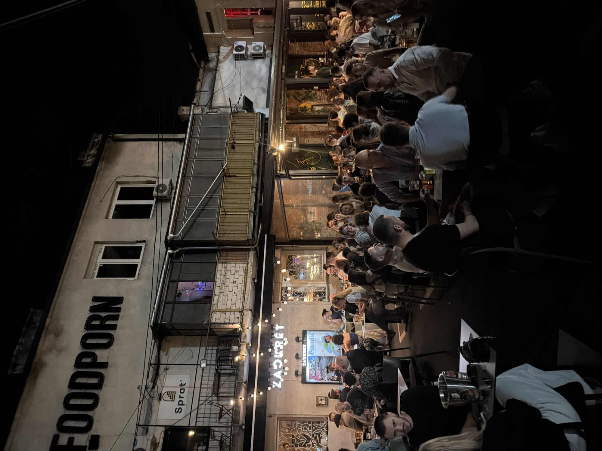 The image shows a night scene at an outdoor seating area of a restaurant or bar in Belgrade, Serbia. The establishment has a large sign reading "FOODPORN" in black lettering on the upper facade of the building. Below this, another sign reads "ZADRZET" in neon. The area is filled with people seated at tables, socializing and eating or drinking. String lights are strung across the outdoor area, illuminating the scene. The architecture appears to be a mix of older buildings with balconies and modern renovations. The overall atmosphere is lively and crowded, typical of a popular urban night spot.
