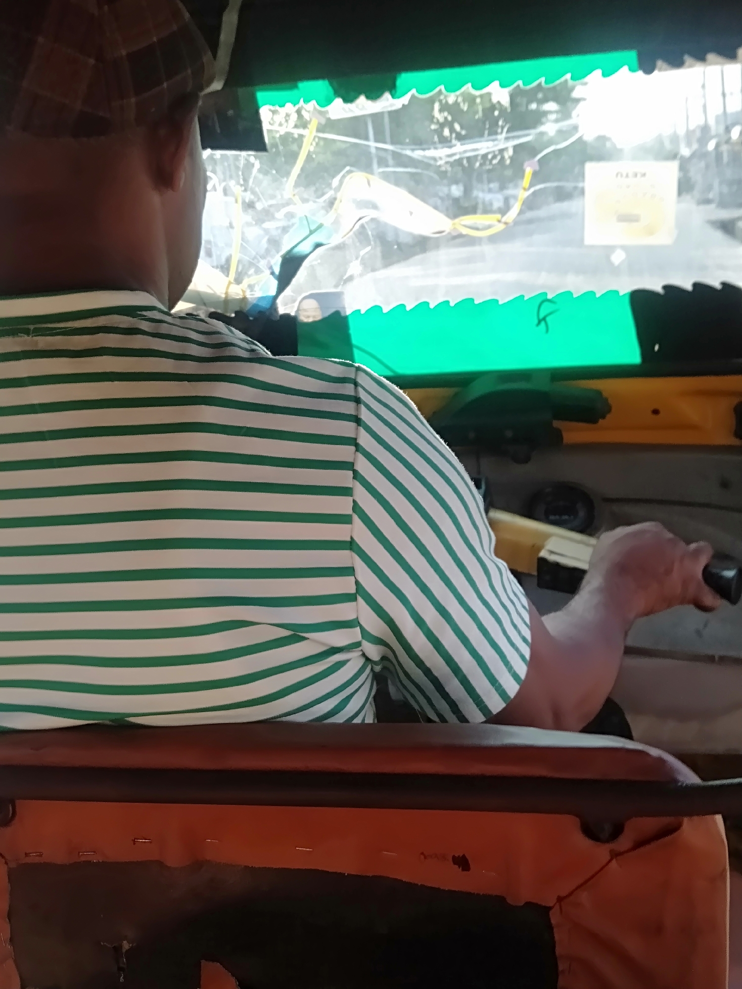 The image depicts a man operating what appears to be an auto-rickshaw (tuk-tuk) from the driver's seat. The man is wearing a hat and a white shirt with green stripes. The windshield of the auto-rickshaw appears damaged, with visible cracks. Outside the vehicle, the street is visible, with buildings and trees suggesting an urban setting. A sticker or sign with the text "KETU" is seen on the dashboard. The environment suggests a possible location in an Asian city given the type of vehicle and general street scene. The exact location within the city of Unknown, Unknown cannot be determined with the given information.