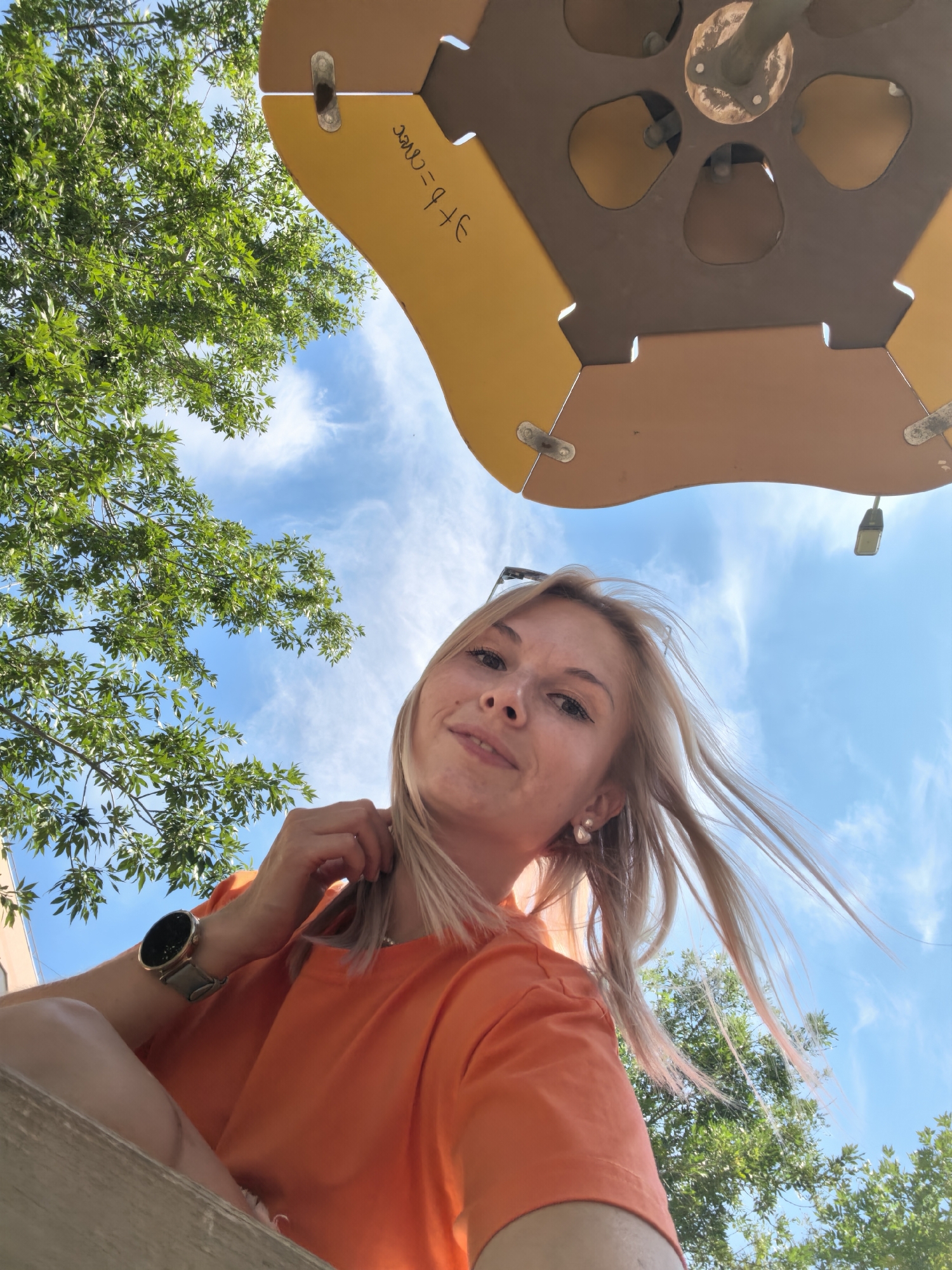 A young woman with blonde hair and an orange t-shirt is captured in a low-angle shot outdoors. She is wearing a watch on her left wrist and earrings. The background features lush green trees against a bright blue sky with scattered clouds. The scene appears to be a sunny day, likely in a park or recreational area. There is a yellow and brown structure overhead, possibly part of a playground or an outdoor seating area. There is no identifiable information or context suggesting the location is in Sterlitamak, Russia.