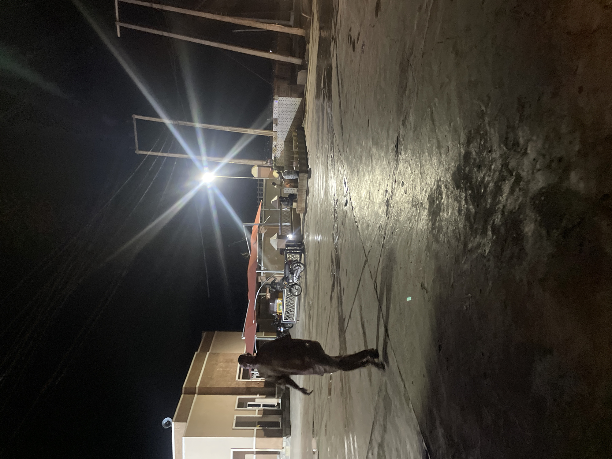 The image shows a person lying on a concrete surface in Hayin Banki, Nigeria. The lighting is poor, but it appears to be nighttime. There is a building in the background, and there are some power lines. The person is wearing dark clothing and appears to be unconscious or dead. There are some motorcycles parked in the background. The ground is wet.