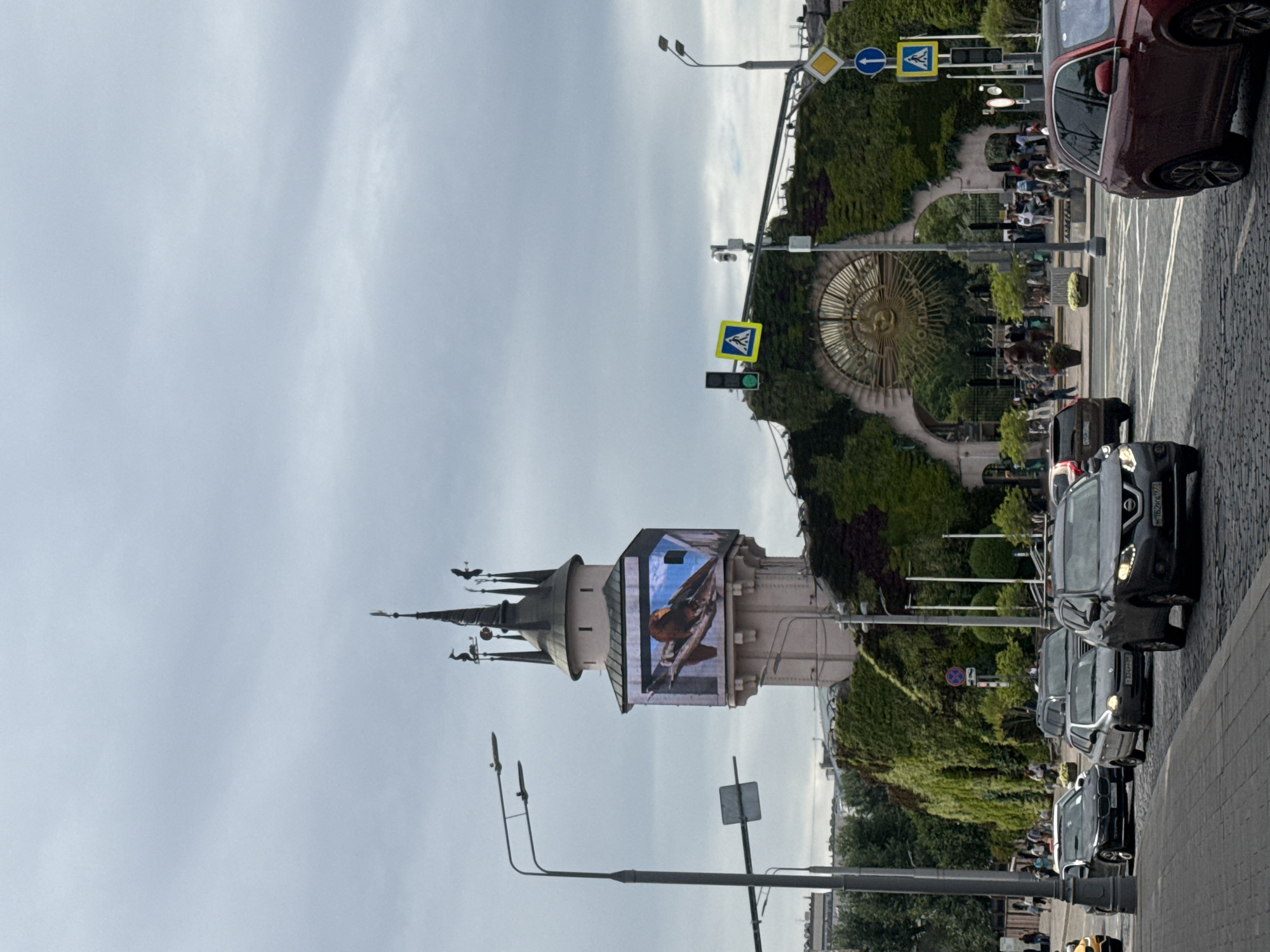 The image shows a street scene in Moscow, Russia, featuring the Moscow State Circus (Bolshoi Circus) on Vernadsky Avenue. In the foreground, several cars are driving on a cobblestone street under a cloudy sky. To the left, a tall lamppost with multiple lights stands prominently. In the background, the distinctive tower of the Moscow State Circus is visible, adorned with a large LED screen displaying an image of a bird. The building's facade is covered in lush greenery, forming an archway with a large, ornate golden sun symbol above the entrance. People are seen walking around the entrance and gathered in front of the circus building. Traffic lights and pedestrian crossing signs are also present, indicating a busy urban environment.