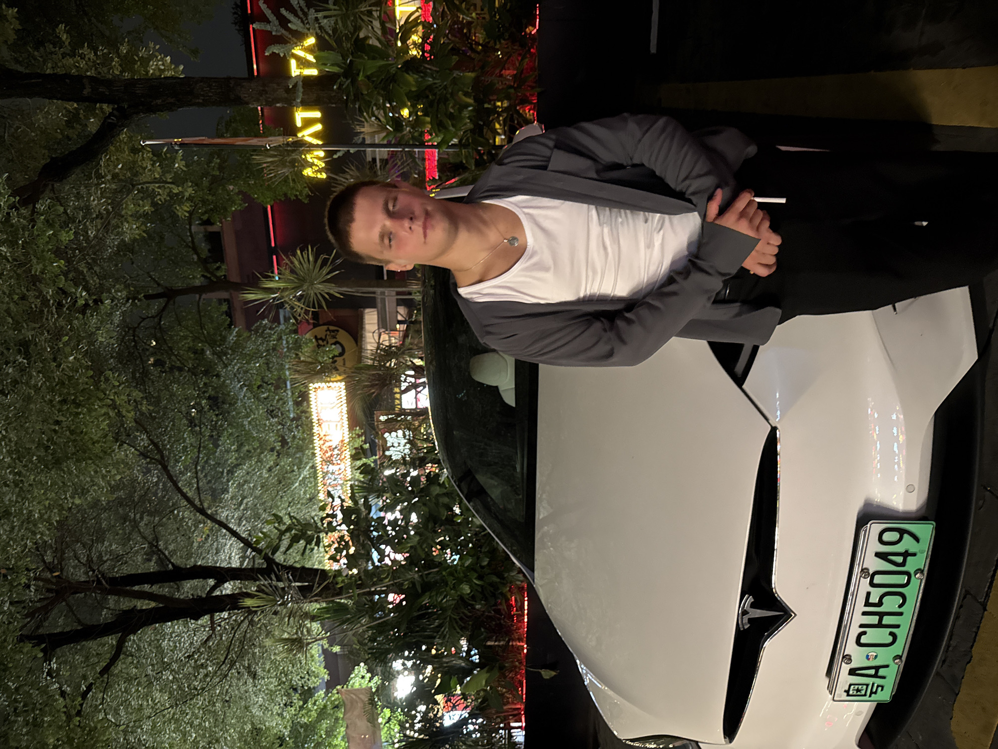 A man is posing on the hood of a white Tesla vehicle in an outdoor environment at night. He is fair-skinned and wearing a white t-shirt, a grey jacket, and a necklace. The car has a green license plate that reads "粤A·CH5049," indicating it is registered in Guangdong Province, China. The background includes trees, streetlights, and blurred signs with neon lighting, one visible sign reads "MYATA". The environment indicates an urban location possibly in Tianhe District, China. The man is looking directly towards the camera with a neutral expression.