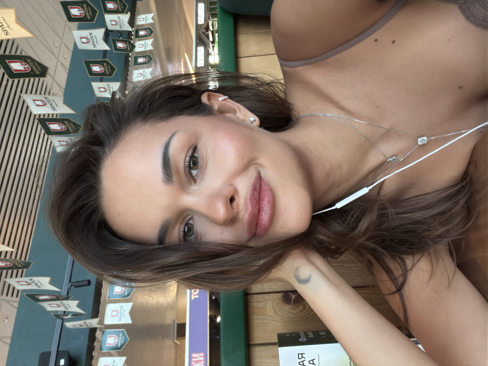 A headshot of a Caucasian woman with long brown hair, light skin, and a gentle smile. She has a mole on her upper chest. She is reclining with her head resting on her hand, which bears a small crescent moon tattoo on the wrist. She is wearing a light brown bra and two delicate silver necklaces. A white earbud cable is visible near her neck.
In the background, there are decorations with "SPATEN" written on them, suggesting the photo was taken in a bar or restaurant. The text "КИ АЯ" and parts of a book title in Russian are partially visible near the woman’s arm. The partial Russian book title is "... Фести... / ГИСТРАЦИЮ /... А." The book may be "ФЕСТИВАЛЯ АРХИТЕКТУРЫ."
The image was taken in Sukhaya, Russia.