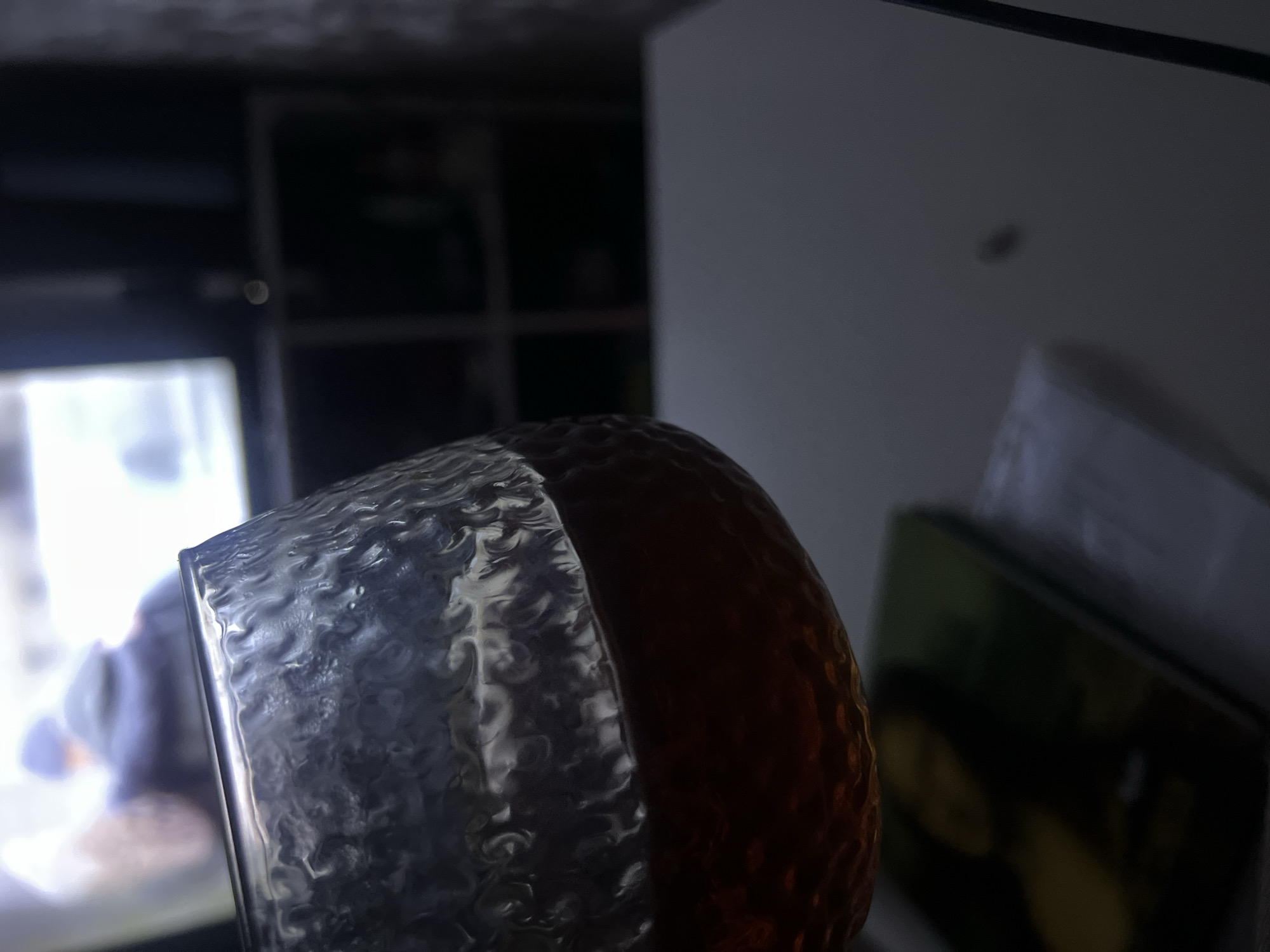 The image shows a close-up of a textured glass or container. The container appears to be partially filled with a dark amber liquid. In the blurred background, a television screen is visible, displaying a person. A shelf or storage unit is situated behind the television. To the right, a white surface, possibly a wall or cabinet, is visible with paperwork nearby. The scene appears to be indoors, possibly in a residential setting, based on the furniture and decor. There is no information regarding the location in the city of Chaikovsky, Russia in the picture.
