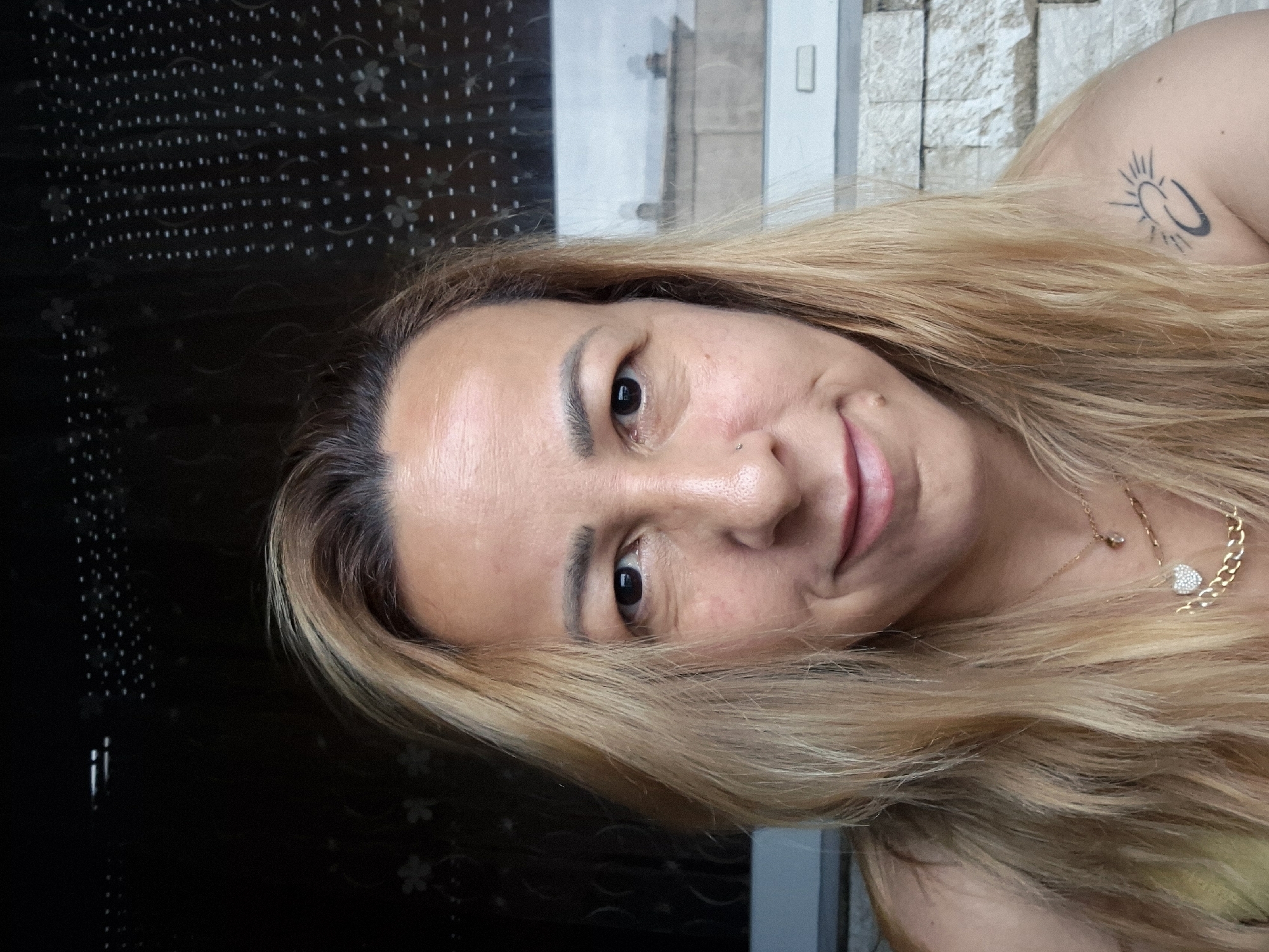 A medium close-up, front-facing portrait captures an adult female with shoulder-length, light blonde hair and a light skin tone, displaying a subtle, closed-mouth smile. Her eyes are dark brown, accentuated by dark eyeliner. Visible fine lines are present around her eyes and forehead, and a small mole is located below the left side of her mouth. A small silver stud pierces her left nostril. She wears multiple gold-colored necklaces, including one with a paved heart-shaped pendant, a thicker chain with chunky links, and another delicate chain with a small dark pendant. On her left shoulder, a black tattoo depicts a stylized sun radiating from a crescent moon, positioned above an abstract linked or infinity symbol. A sliver of a light-colored top is visible on her right shoulder.
The background consists of a reflective surface, indicating a window. The left side of the background is dark, reflecting an interior with faint white dots arranged in vertical lines and subtle floral patterns. The right side reflects an outdoor scene, revealing blurred architectural elements including a light-colored, stacked stone wall in the lower portion and distant buildings with a pitched roof and two small dome-like structures reflected above, under a bright, possibly overcast, sky. A white horizontal window frame is visible in the lower right background. The subject is positioned indoors, looking directly into the camera. The location is in the city of Unknown, Unknown.