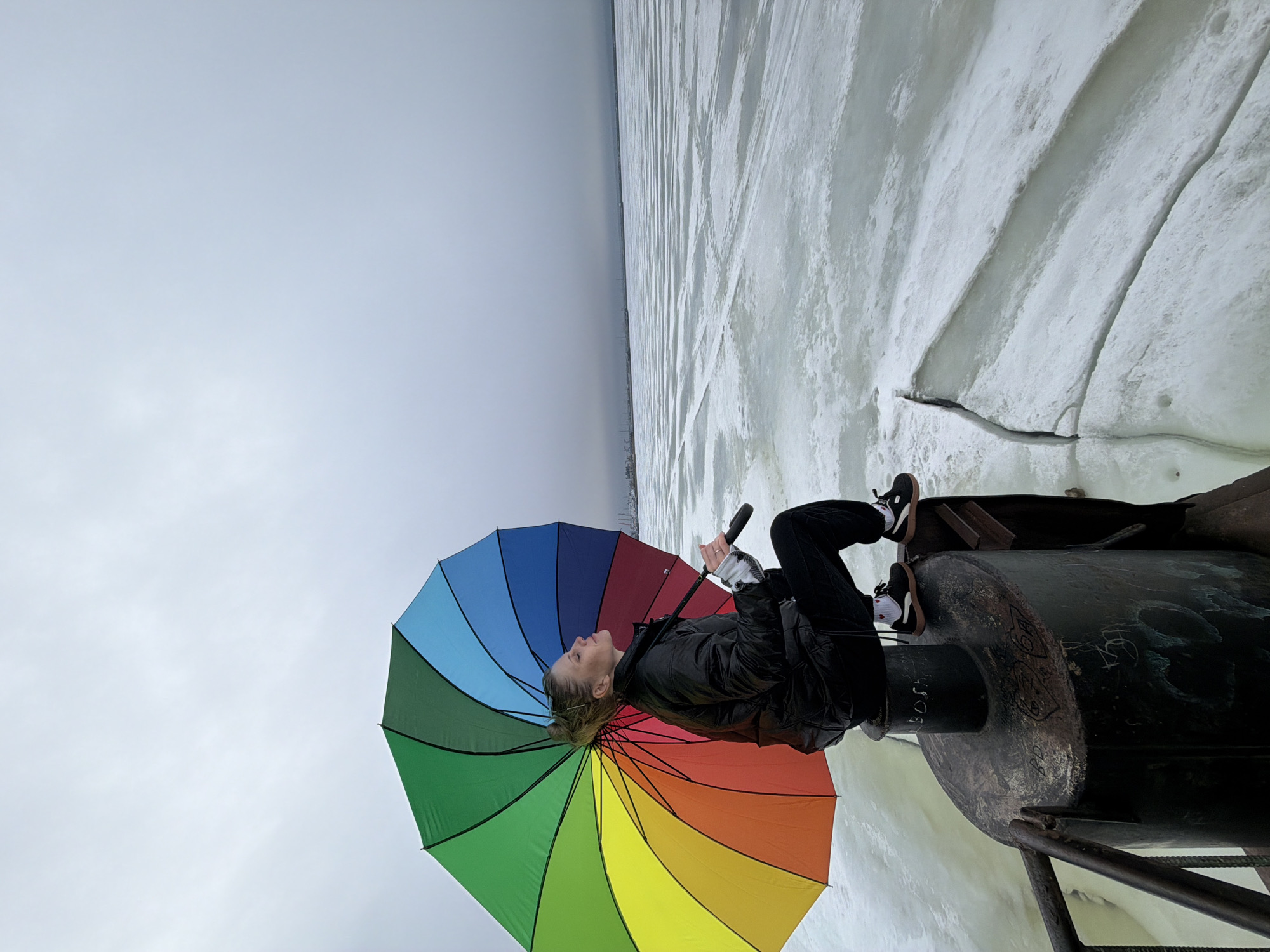A person stands atop a cylindrical metal structure adjacent to a frozen body of water under an overcast sky. The location is Chaikovsky, Russia, evidenced by graffiti visible on the metal structure. The person, dressed in black pants and jacket with brown and black shoes, holds a closed, multicolored umbrella. The background consists of a flat, frozen surface with streaks indicating wind or ice movement, and a gray sky above. The cylindrical structure shows signs of rust and weathering.