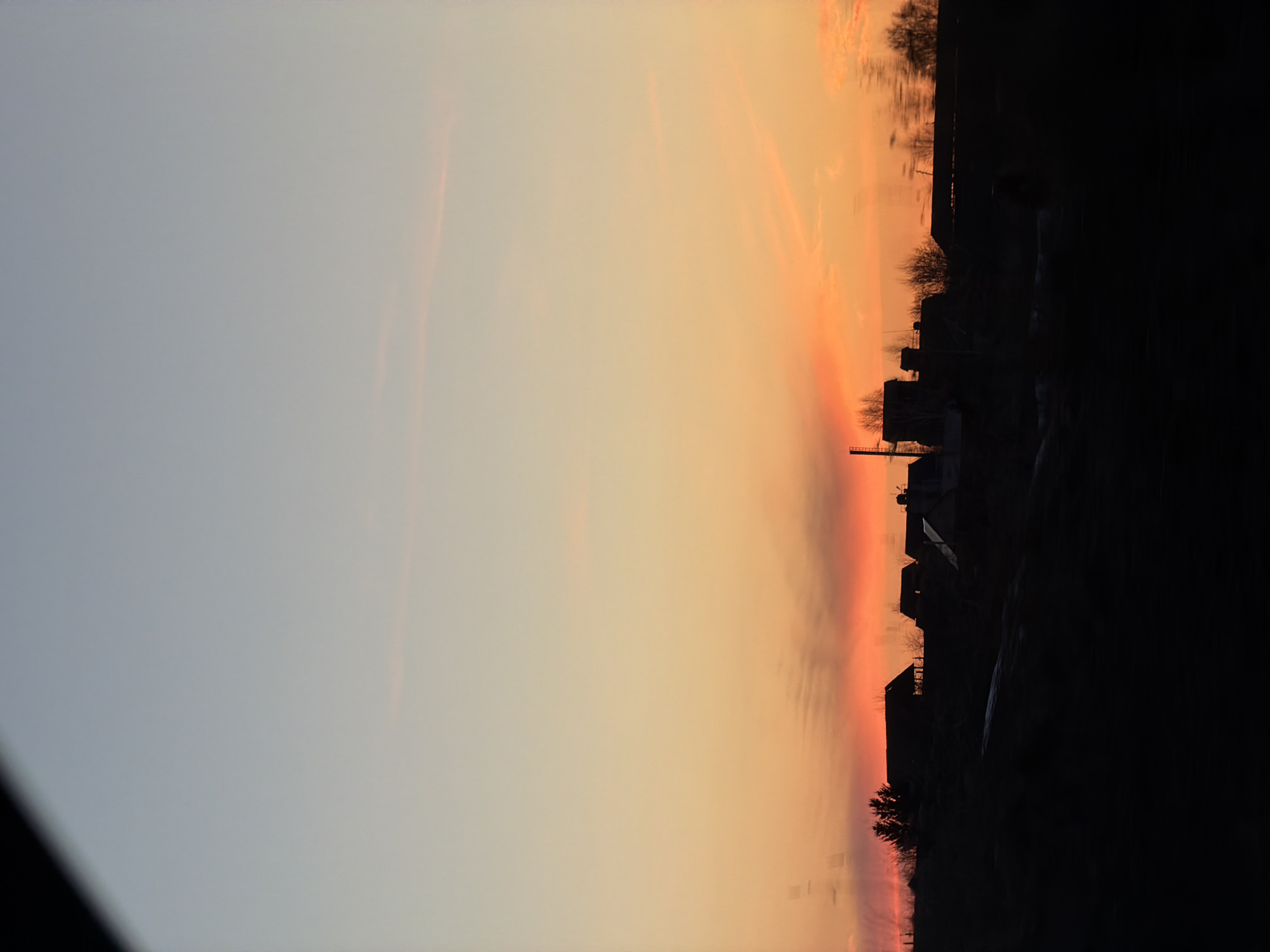 The image depicts a sunset over a location in Gory, Russia. The sky transitions from light gray to orange and red. Silhouetted against the sky are houses and trees. The lower right portion of the image is almost entirely black, obscuring details of the foreground.