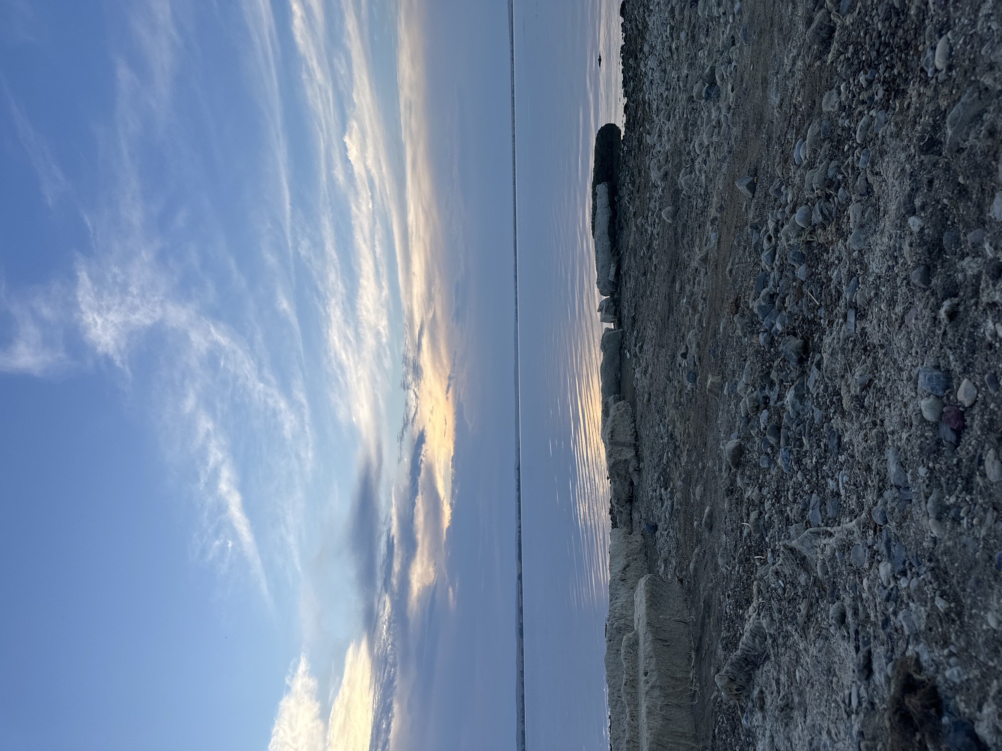 The image depicts a serene natural landscape, likely a body of water near Energetik, Russia. The sky is a soft blue, transitioning to lighter hues near the horizon, with scattered wispy white clouds. The water is calm and reflects the sky, with subtle ripples creating a textured surface. Along the shoreline, the ground is covered with pebbles and rocks of various sizes, mixed with some dry, fibrous material resembling hay bales. Large, light-colored rocks or concrete blocks are positioned near the water's edge. In the distance, a faint line of land is visible on the horizon. The scene appears to be captured during either sunrise or sunset, given the soft lighting and pastel colors in the sky and on the water. There are no people or animals visible in the frame. The environment suggests a quiet, natural setting.