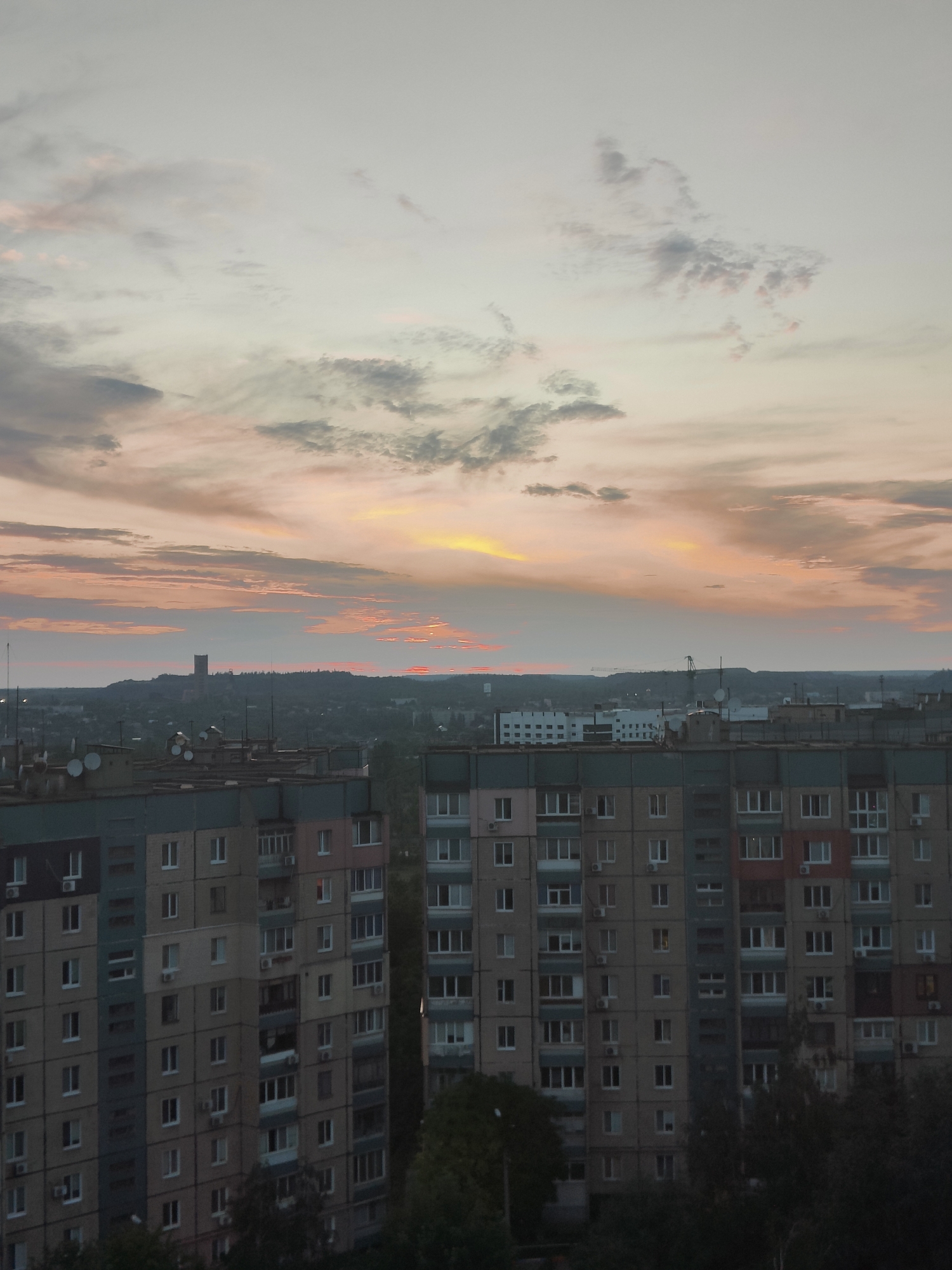 The image shows a cityscape at sunset in Kryvyi Rih, Ukraine. In the foreground, there are several multi-story apartment buildings with a mix of visible windows, balconies, and air conditioning units. The buildings appear to be constructed from concrete and brick, with various colored facades. On the rooftops of some buildings, satellite dishes are visible. In the middle ground, the city extends further, with more buildings and some trees. A distant hill or elevated area is visible on the horizon, topped with a tall, rectangular structure, possibly a tower or industrial building. The sky is filled with dramatic clouds, painted with hues of orange, yellow, and pink from the setting sun, with lighter gray clouds scattered throughout. The overall impression is a view of an urban environment during twilight.
