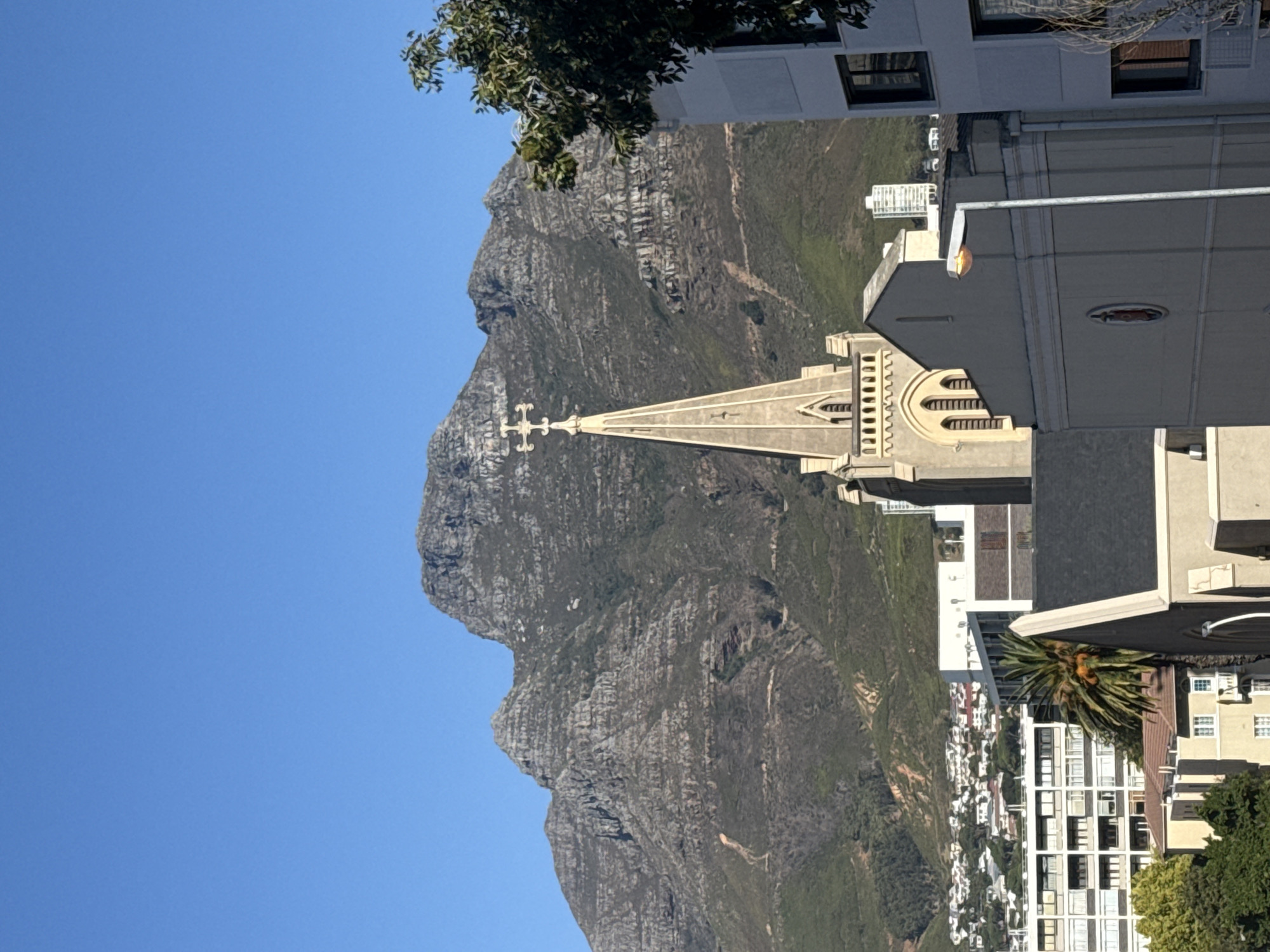 The image presents an urban landscape in Cape Town, South Africa, featuring Table Mountain under a clear blue sky. Table Mountain dominates the background, exhibiting rugged, rocky terrain with prominent horizontal strata and patches of green vegetation. Visible tracks or erosion patterns are present on its lower slopes. In the mid-ground, a stone-colored church spire with a cross at its apex rises prominently, characterized by arched windows and decorative elements. Surrounding the church are various multi-story buildings, including modern white structures with dark window frames and older buildings with different rooflines. A green tree branch is visible in the upper right, and a palm tree is situated among the buildings. A grey street lamp with an illuminated yellow globe stands in the lower-right mid-ground, obscuring part of a building. The scene is brightly lit by natural light. No individuals, explicit actions, or direct interactions are visible.
