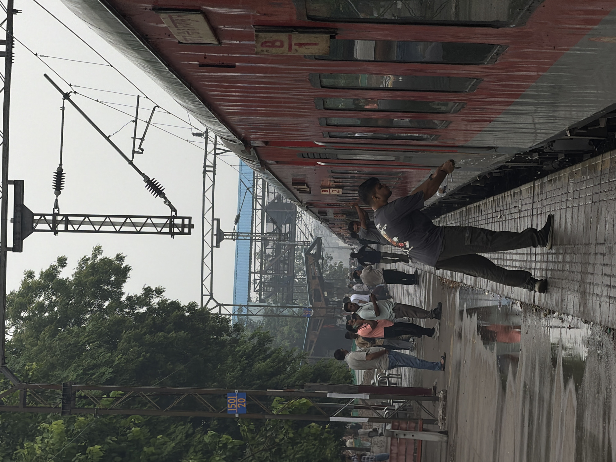 The image depicts a scene at a train station in Ballabgarh, India. A red and grey passenger train is stopped at the platform. The platform is wet, indicating recent rain, with puddles reflecting the overhead electrical wires and parts of the train. Several individuals are present on the platform. On the left, a group of men are standing, some appearing to be waiting. Further down the platform, near the train, a man in a dark grey t-shirt and grey cargo pants is interacting with the train door, possibly closing or opening it. Another person is partially visible on the right side of the frame, also near the train. In the background, a railway overpass structure and trees are visible. A blue sign with white text "1501 / 20" is mounted on a pole to the left of the train. The overall atmosphere is that of a typical Indian railway station, possibly during or after rainfall.