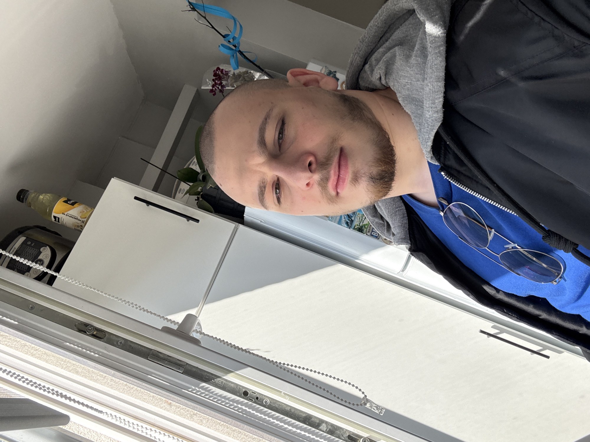 A man with a shaved head and a beard is shown from the chest up. He is wearing a blue shirt, a gray hoodie, and a black jacket. Sunglasses are on his shirt. The background is a window with closed blinds and white cabinets. On top of the cabinets are a plant and a bottle of oil. The location is likely indoors and, as per the prompt, potentially in Stara Zagora, Bulgaria, though there is no visual information in the image to independently verify this.