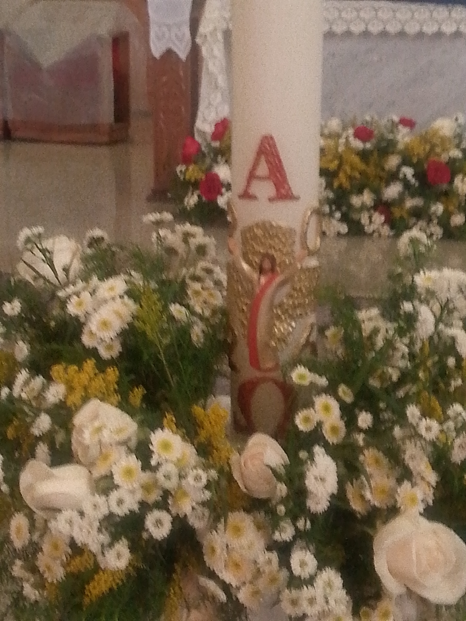 A tall, white candle with a red "A" and a depiction of Jesus Christ with golden wings is centered among white and yellow flowers. The scene appears to be inside a church or religious setting, based on the altar and white lace covering visible in the background. Red flowers are scattered among the background flower arrangements. The location is Caracas, Venezuela, although nothing definitively places the scene in that city within the image itself.