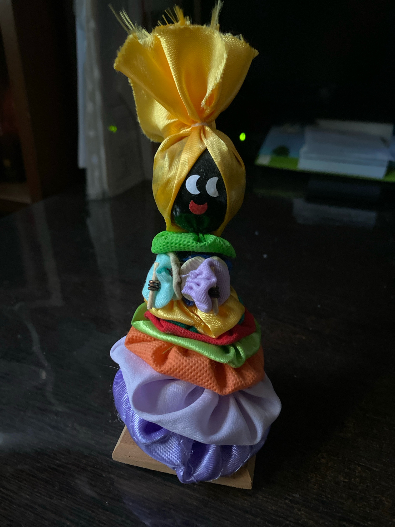 A handmade doll crafted from fabric scraps stands on a dark, reflective surface, likely a table. The doll has a black head with white eyes and a red smile, topped with a yellow fabric headpiece that is gathered and tied. Its body is composed of layers of colorful, ruffled fabric, including green, blue, red, orange, light purple, and dark purple. A small wooden base supports the doll. The background is dark and slightly blurred, with hints of indoor lighting and objects, possibly in a room in Ivanteyevka, Russia.