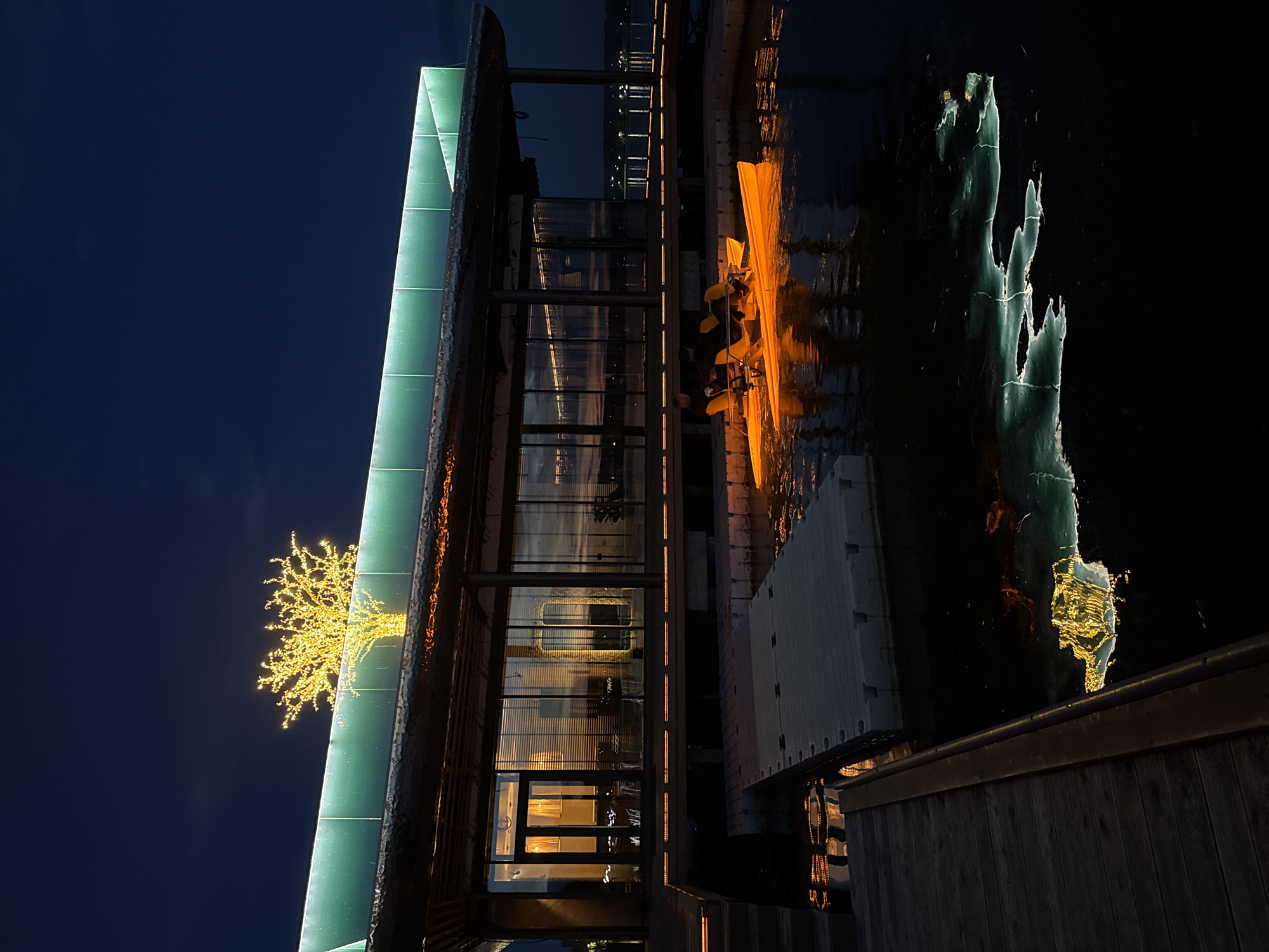The image captures a nighttime scene at a waterfront establishment in Votkinsk, Russia. The main building features large glass windows reflecting the interior lights and the dark blue sky. A prominent illuminated tree, possibly a decorative installation, sits atop the building's green-lit roofline. In the foreground, a wooden dock leads to a floating platform on the water. A bright yellow pedal boat with two occupants is moored to the platform, its lights casting an orange glow on the water. The dark water reflects the lights from the building and the pedal boat, creating abstract patterns. The overall atmosphere is serene and illuminated by artificial light against the night sky.