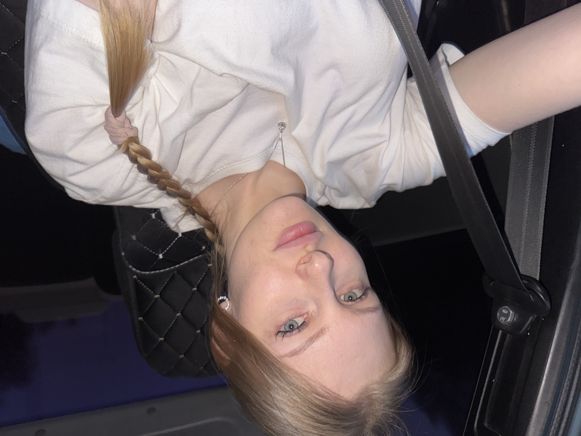 A young, fair-skinned woman with blonde hair, partially braided and secured with a pink scrunchie, is wearing a white long-sleeved shirt and a seatbelt. She is positioned in a vehicle, likely a car, with a quilted black seat cover visible. The environment is dark, suggesting nighttime or an enclosed space, and the location might be Perm, Russia.