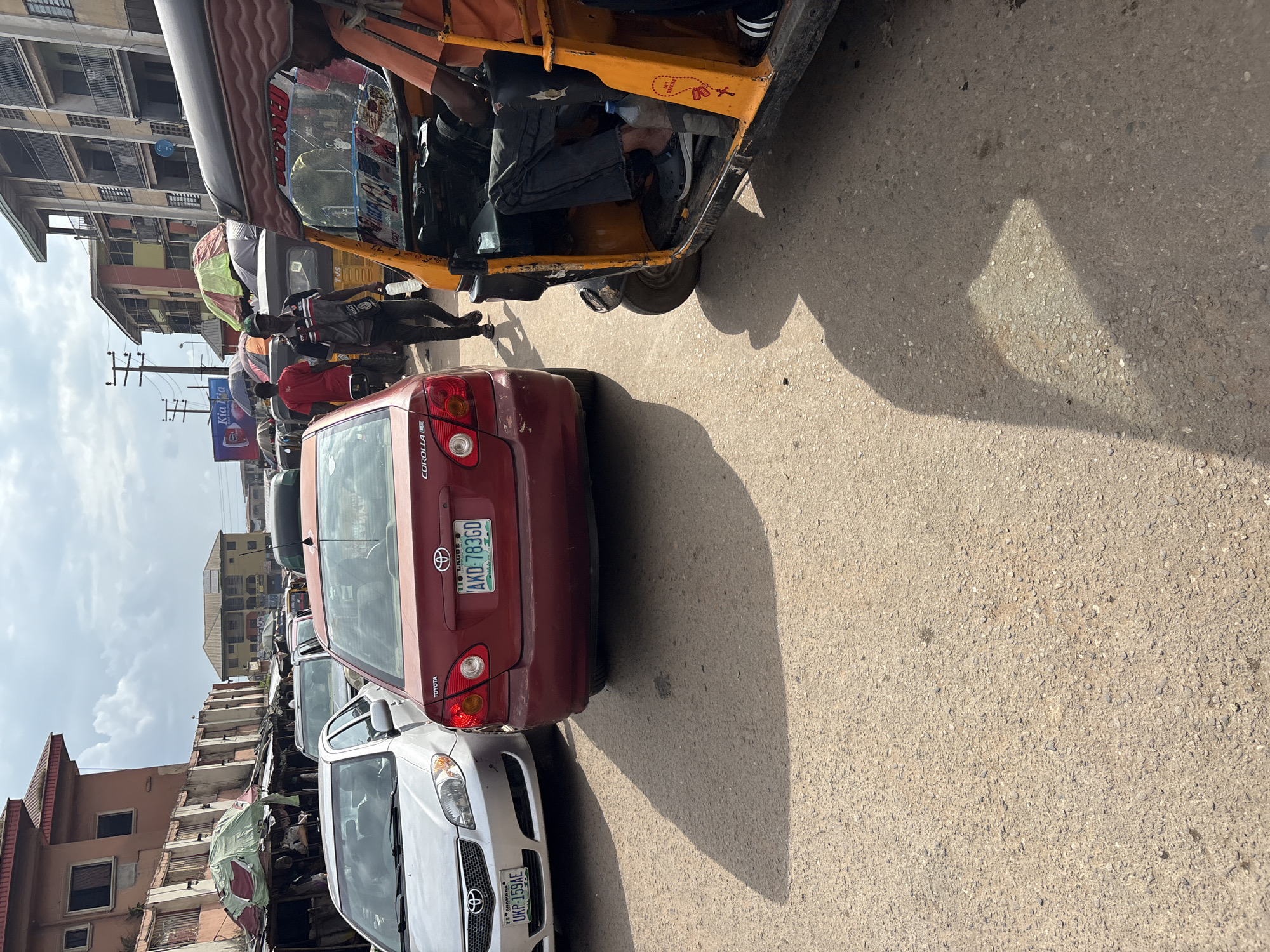 A street scene in Onitsha, Nigeria, depicts a busy thoroughfare with multiple vehicles, including cars and a three-wheeled auto-rickshaw. In the foreground, a red Toyota Corolla is parked, its rear license plate reading "AKD 783GD". To its left, a silver sedan with a license plate "UKP-159AE" is also visible. A yellow auto-rickshaw with a driver wearing an orange shirt and dark pants occupies the right side of the frame. People are seen walking on the roadside, some under makeshift stalls selling various goods. Buildings line the street, appearing to be commercial or residential structures. The sky is partly cloudy.