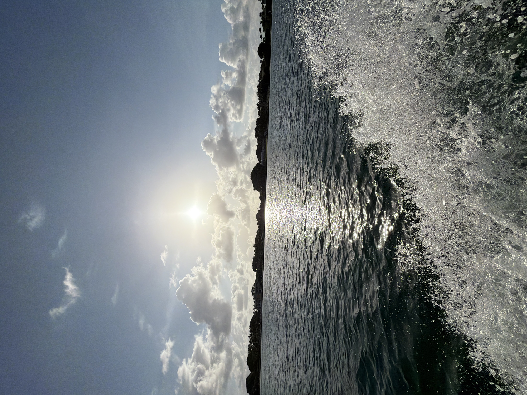 The image shows a scene from the water, likely a bay or inlet, near North Miami, Florida. The view is oriented vertically, with the sky and clouds on the left, and the water on the right. The sun is visible through the clouds, creating a bright glare. A landmass with trees and some buildings is situated between the water and the sky. The water is dark and rippled, with sunlight reflecting off the surface. On the right edge of the water, there is a splash or wave. It's unclear if this is caused by a boat moving through the water or natural wave action. No people are visible.