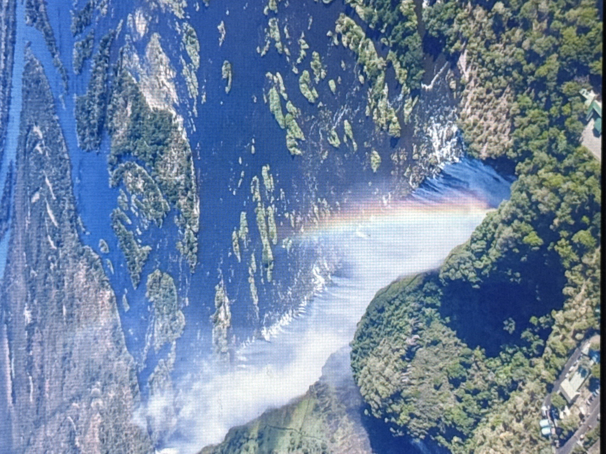 This is an aerial view of Victoria Falls, a waterfall on the Zambezi River in southern Africa, located on the border between Zambia and Zimbabwe. The image shows the immense cascade of water plunging into a gorge, creating a thick mist that refracts sunlight into a prominent rainbow. The surrounding landscape is densely forested with lush green trees, and the river above the falls is wide and appears to be dotted with numerous small islands. The water is a deep blue, contrasting with the white spray of the falls and the vibrant green of the vegetation. In the lower left portion of the image, some man-made structures and roads can be seen, suggesting the presence of tourist facilities near the falls. The scene is bathed in sunlight, highlighting the natural beauty and grandeur of this natural wonder.
