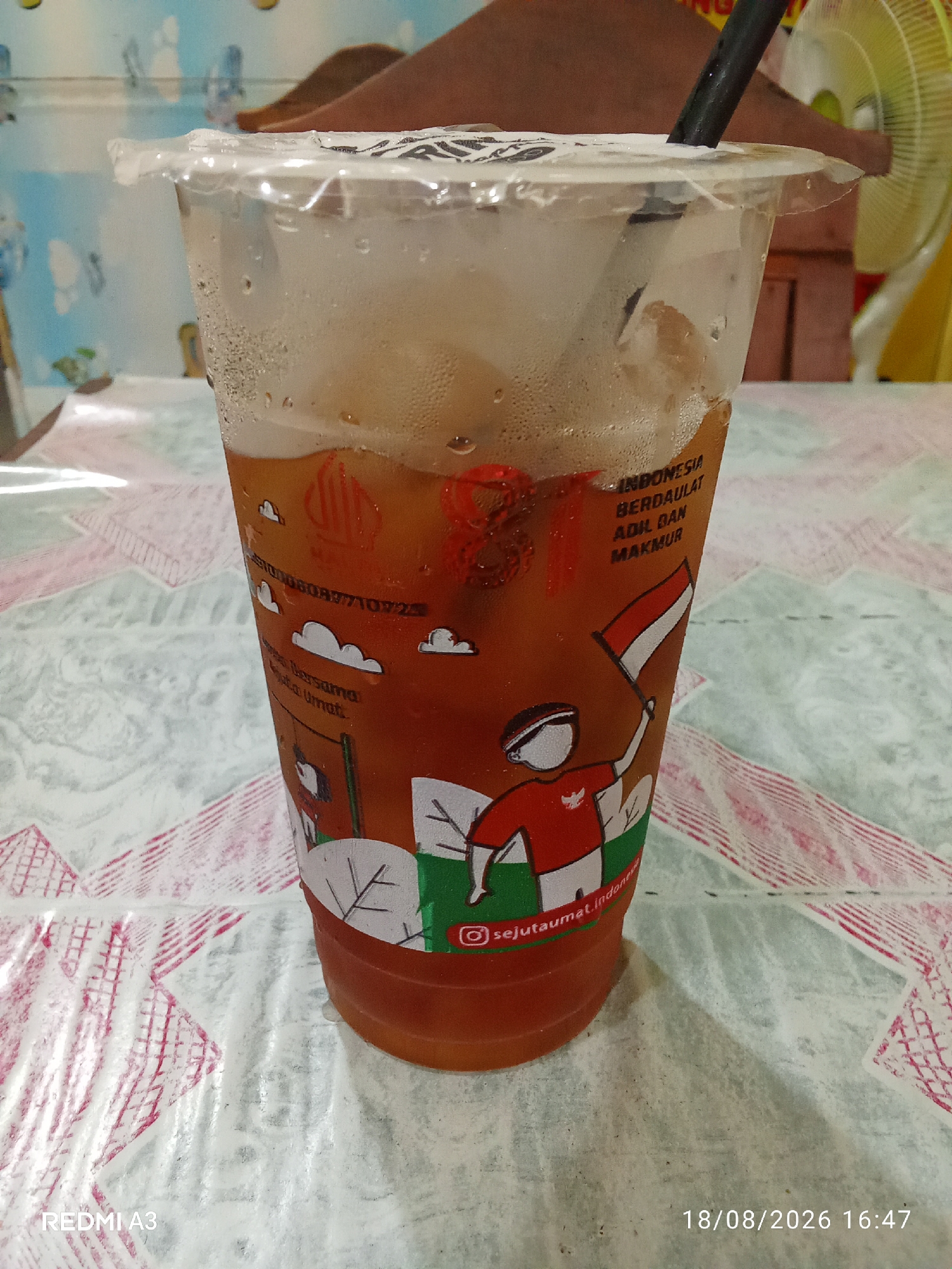 A disposable plastic cup filled with iced tea and ice is positioned on a table covered with a patterned tablecloth. The cup has condensation on its exterior and a clear plastic lid with a black straw inserted. The cup is decorated with graphics that appear to be related to Indonesian national pride, including text that reads "INDONESIA BERDAULAT ADIL DAN MAKMUR" (Indonesia Sovereign, Just and Prosperous) and images of people holding Indonesian flags. A small Instagram logo and the handle "@sejutaimat.indone" are visible on the cup. The background shows a blurred environment with a fan and a table. The bottom left corner of the image displays the text "REDMI A3", and the bottom right corner shows the date and time "18/08/2026 16:47". The context of Banaran, Indonesia, is not visually present in the image itself.