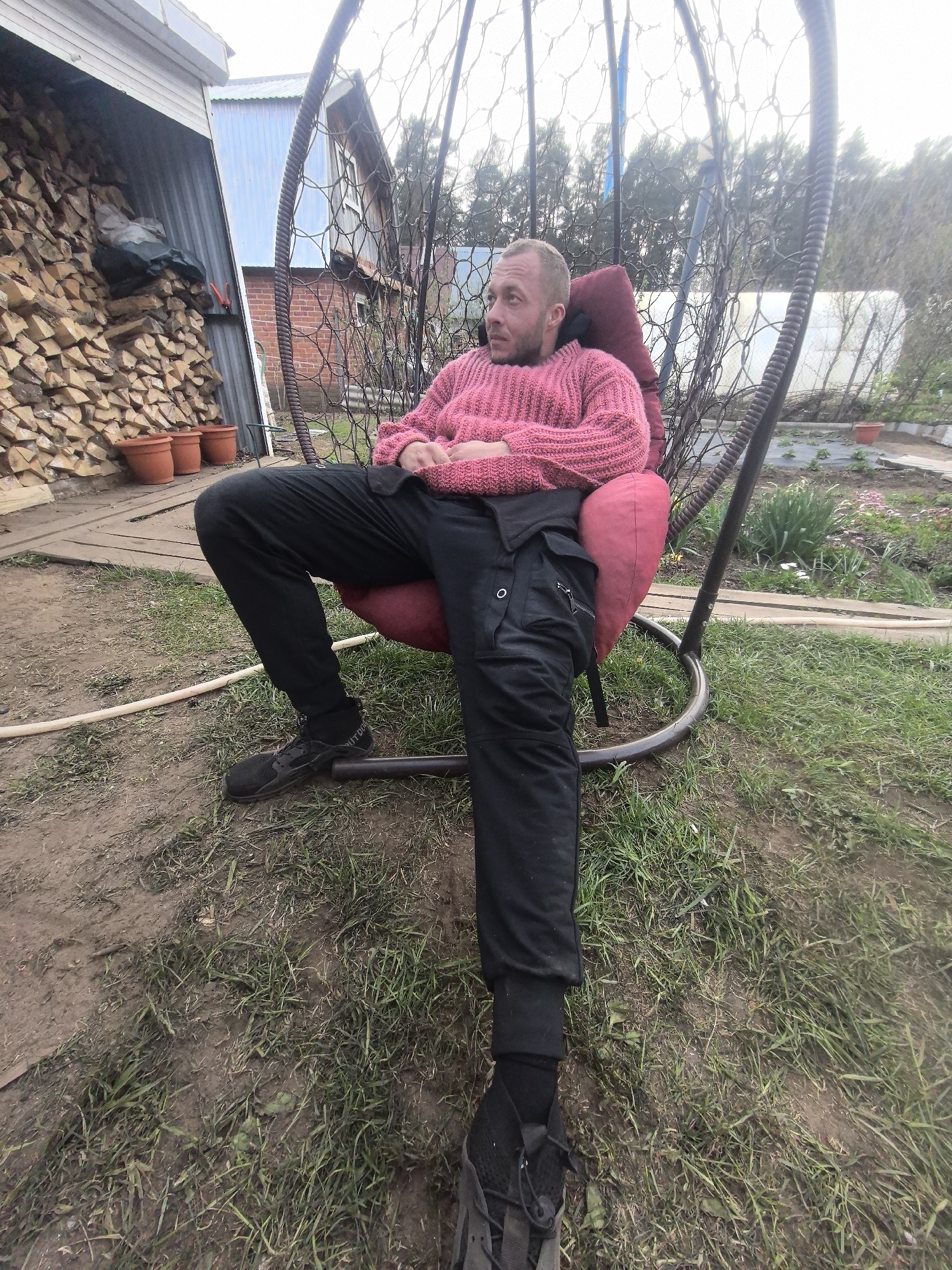 A man sits in a hanging egg chair on a lawn. He is wearing a pink sweater and black pants, with black sneakers. He is fair-skinned with short hair and a beard. The chair is red and gray. Behind the chair is a garden, and in the background are several buildings, one with exposed brick. A large stack of firewood is visible in the background as well, under a corrugated metal shelter. The ground is dirt and grass. This photo was taken outdoors, presumably during the day. The specific location, Noviy, Russia, is inferred from the query, but there are no distinct features in the image to confirm it.